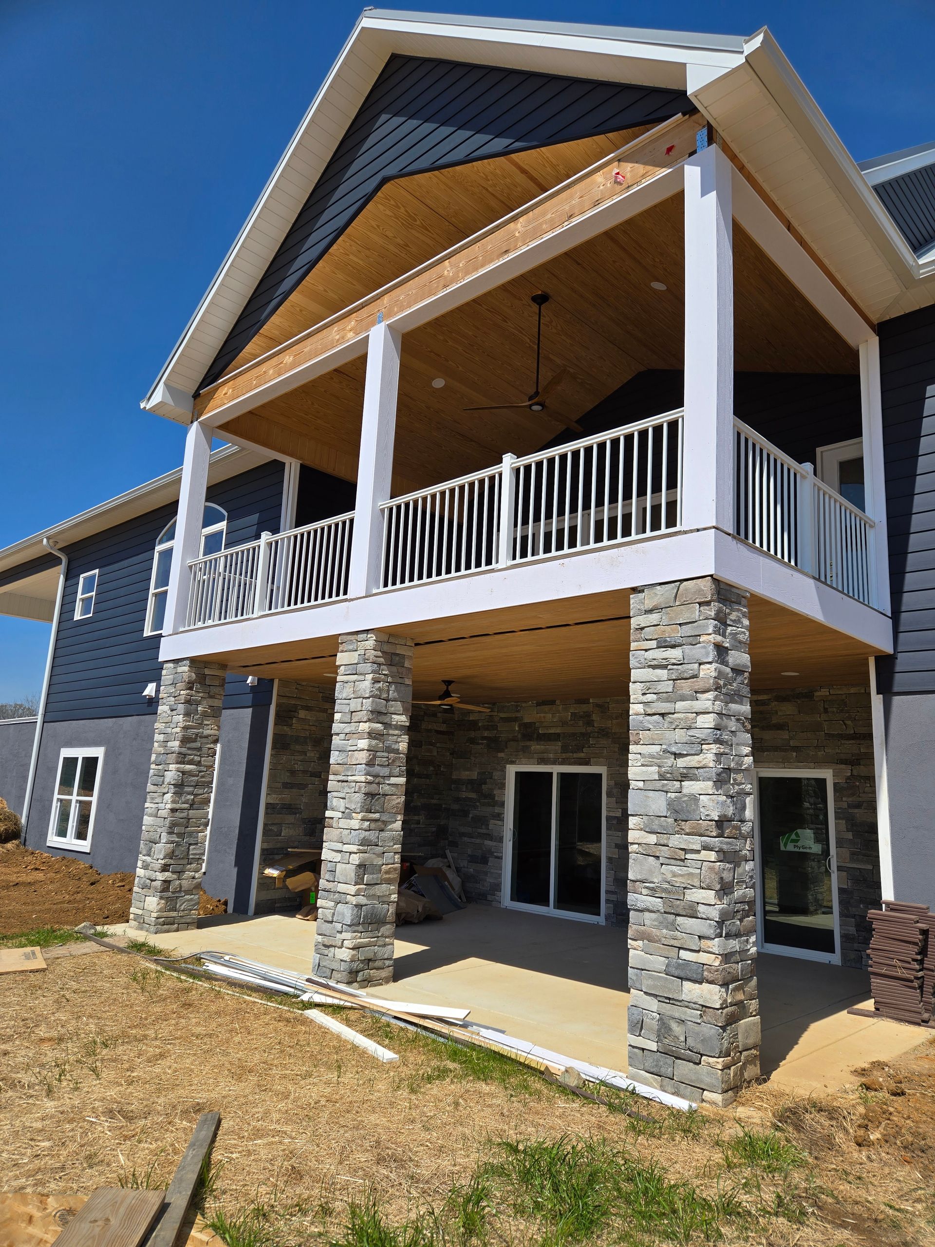 Two-story house with stone columns, white balcony railings, and a covered porch under construction.