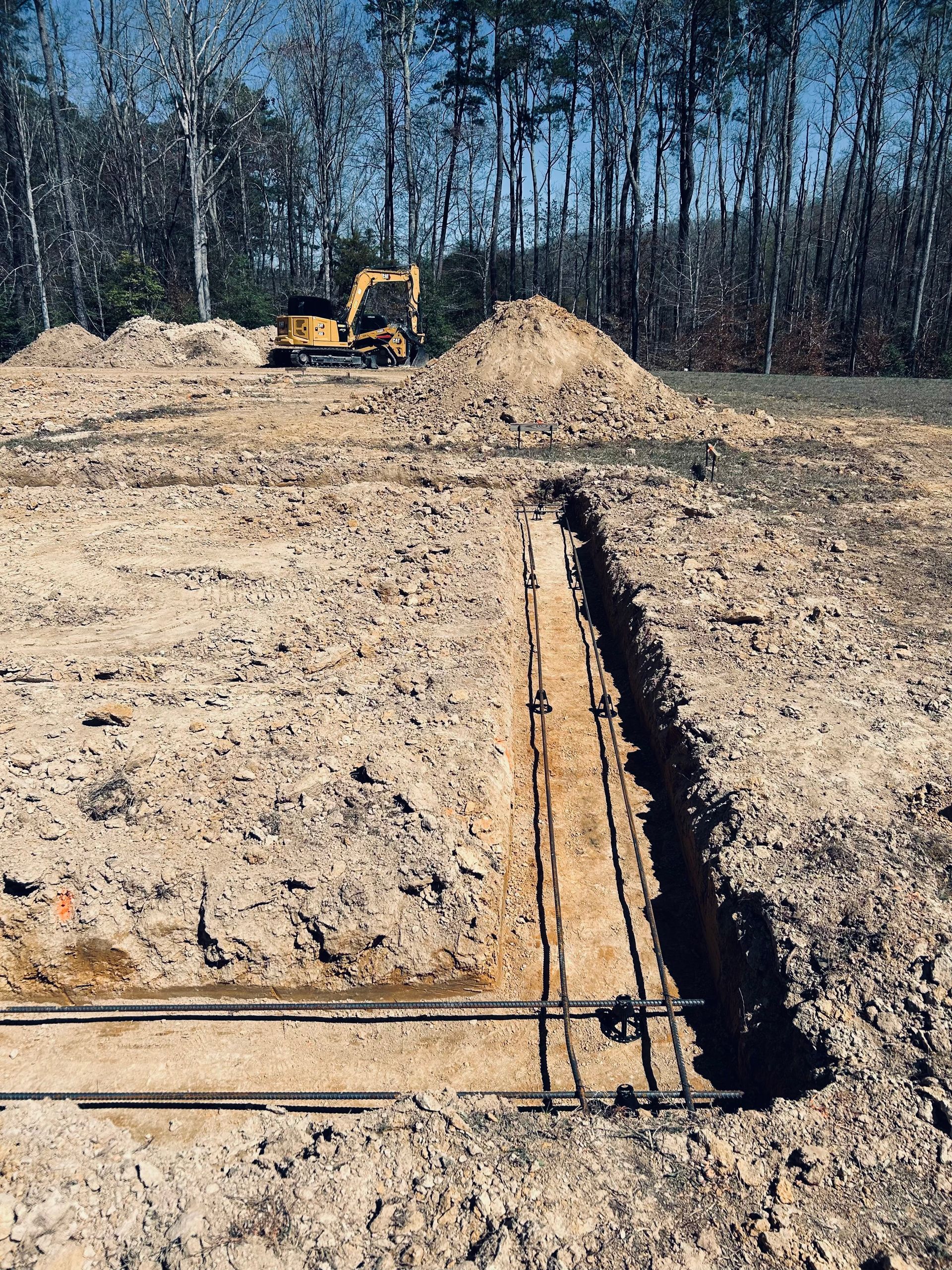 Excavator beside a trench at a construction site with stacked pipes and a dirt mound under a tree line