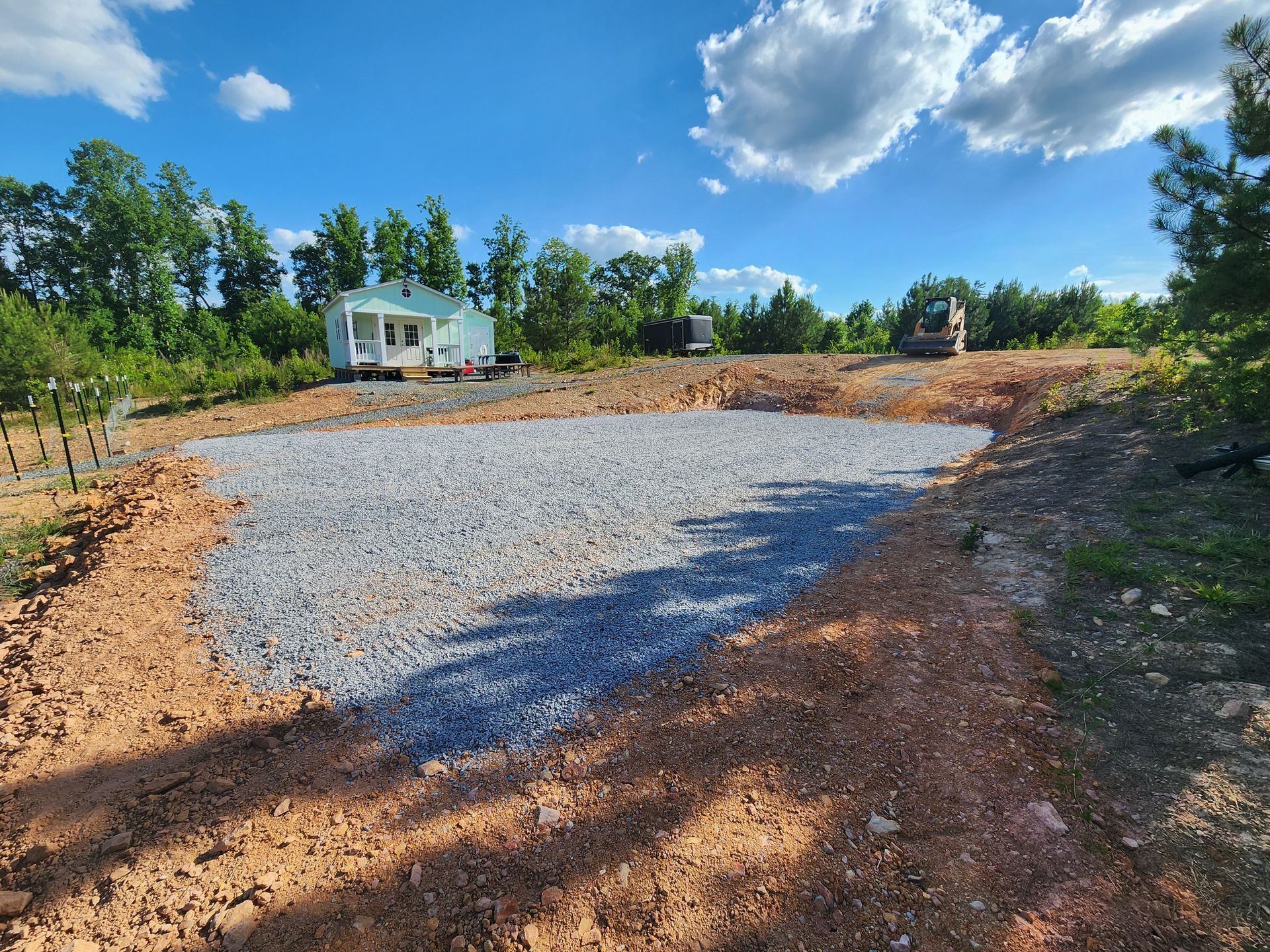 Gravel driveway under construction beside a white tiny house, with trees and blue sky in the background