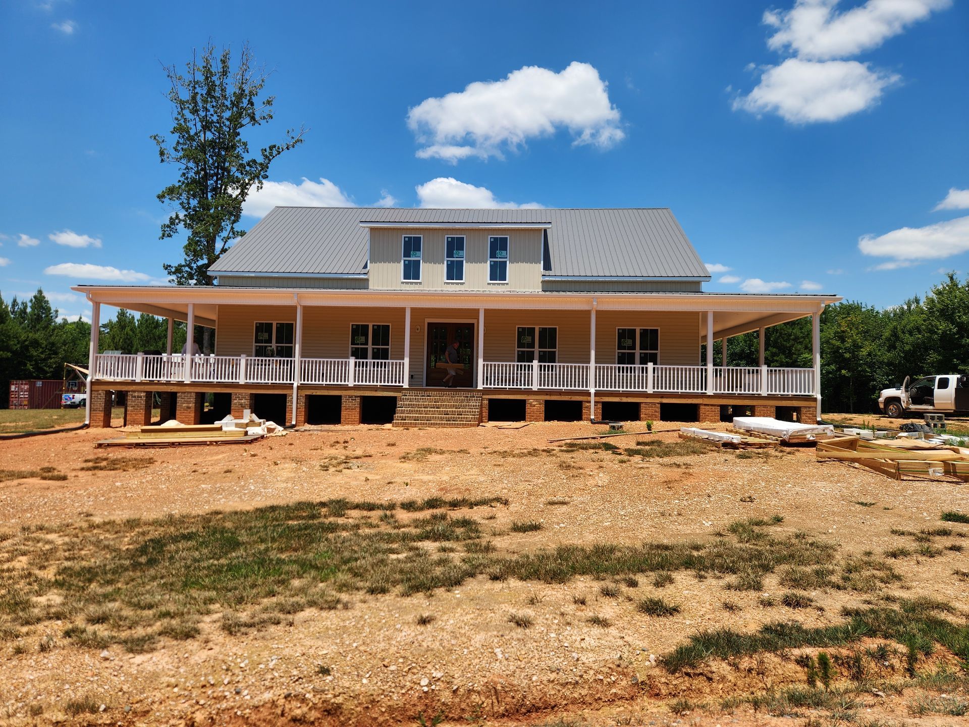 Large unfinished house with a wraparound porch on a dirt lot under a blue sky