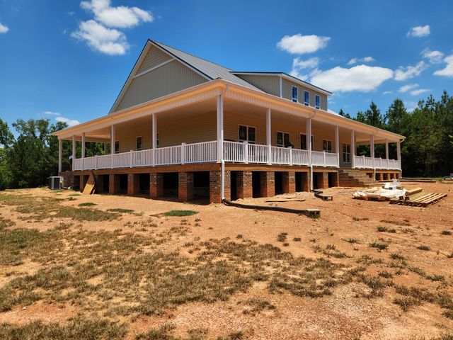 Large white elevated house with wraparound porch under construction on a sandy lot under a blue sky
