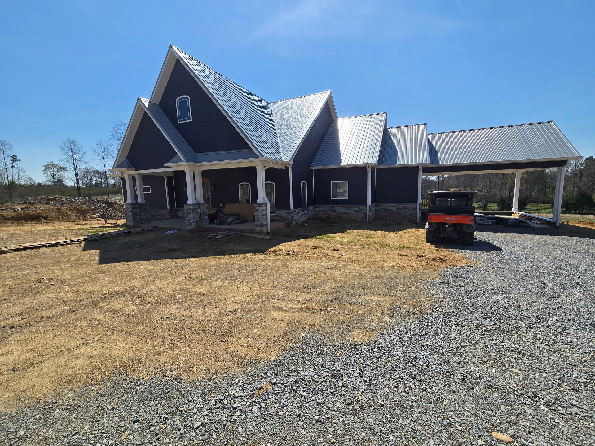 Gray house with steep roof and porch, gravel driveway, and a red truck under a carport on a sunny day