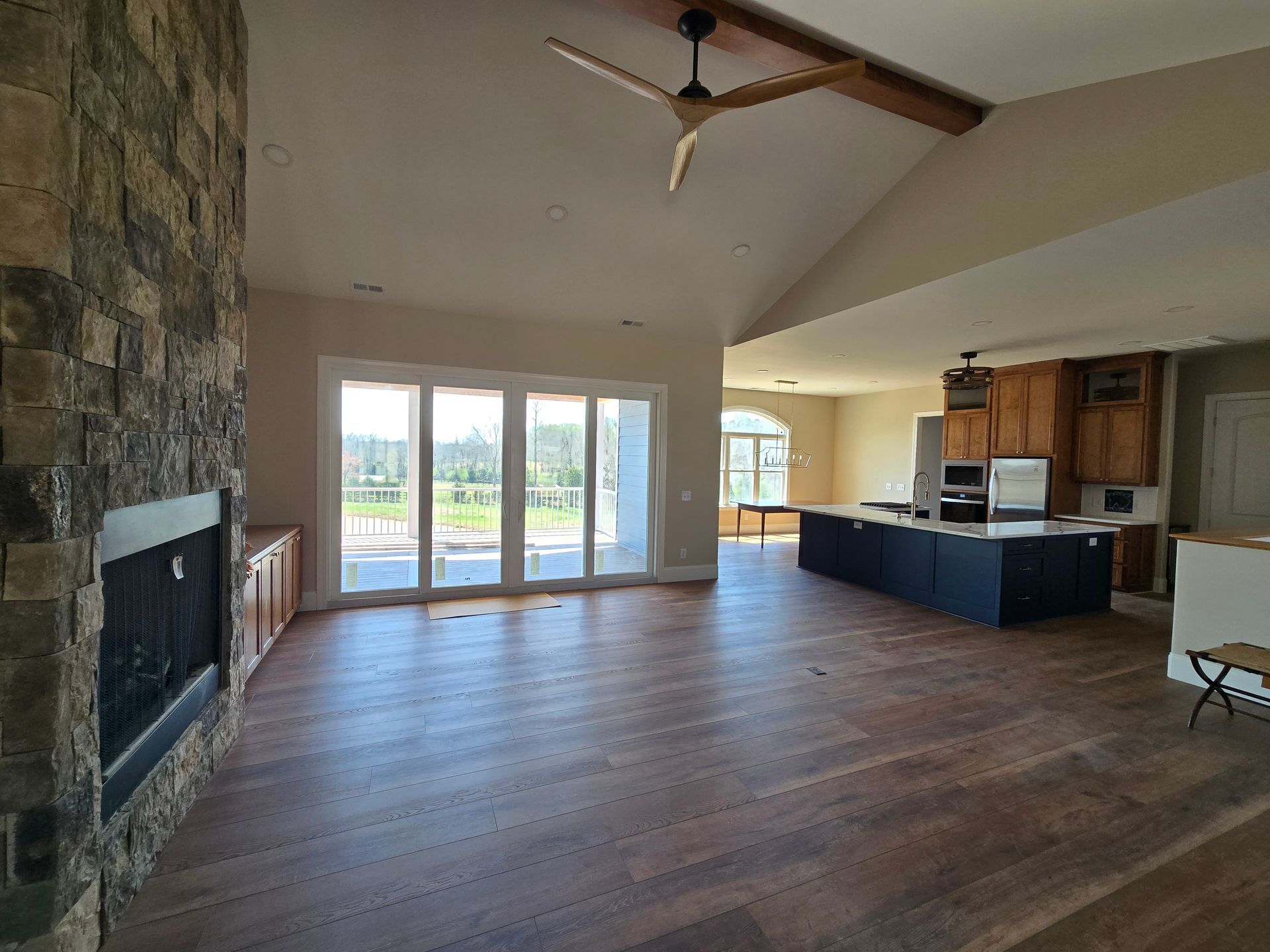 Empty living room with stone fireplace, large windows, and open kitchen with dark cabinets