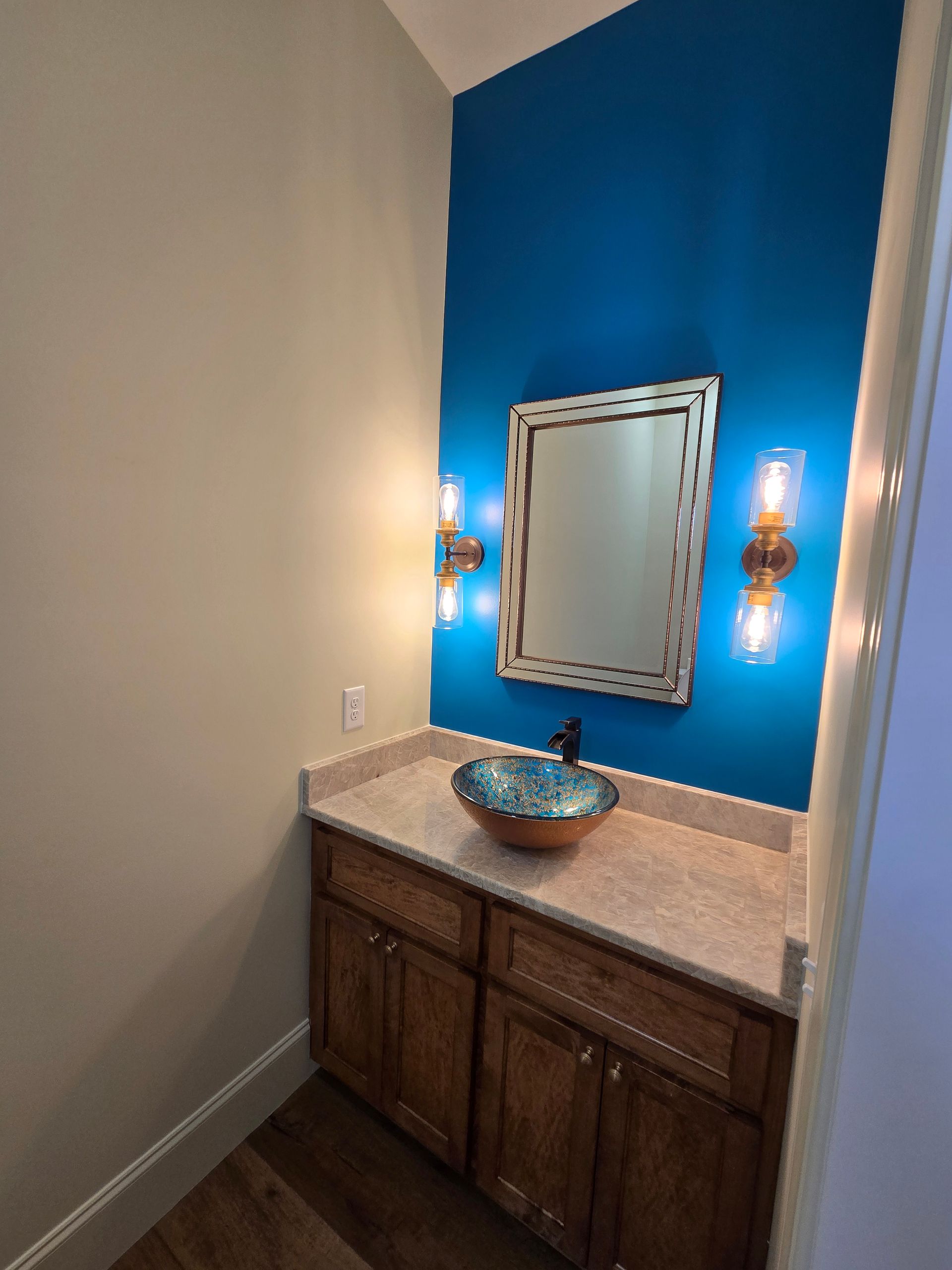 Bathroom vanity with blue accent wall, mirror, sconces, and a stone countertop with bowl sink