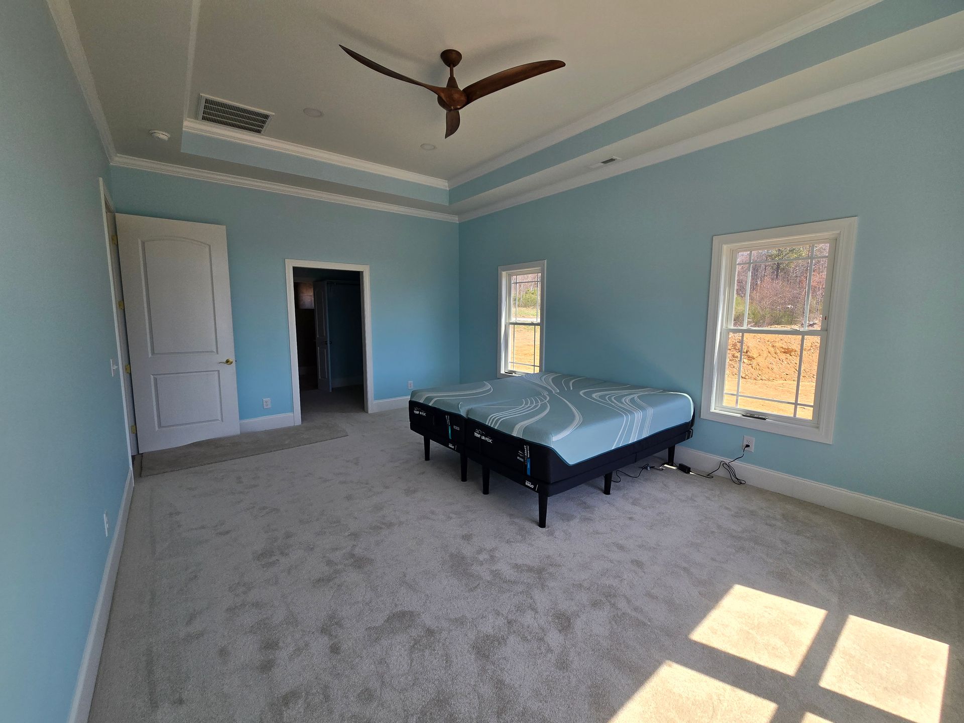 Spacious light blue bedroom with gray carpet, bed, ceiling fan, and sunlight through windows