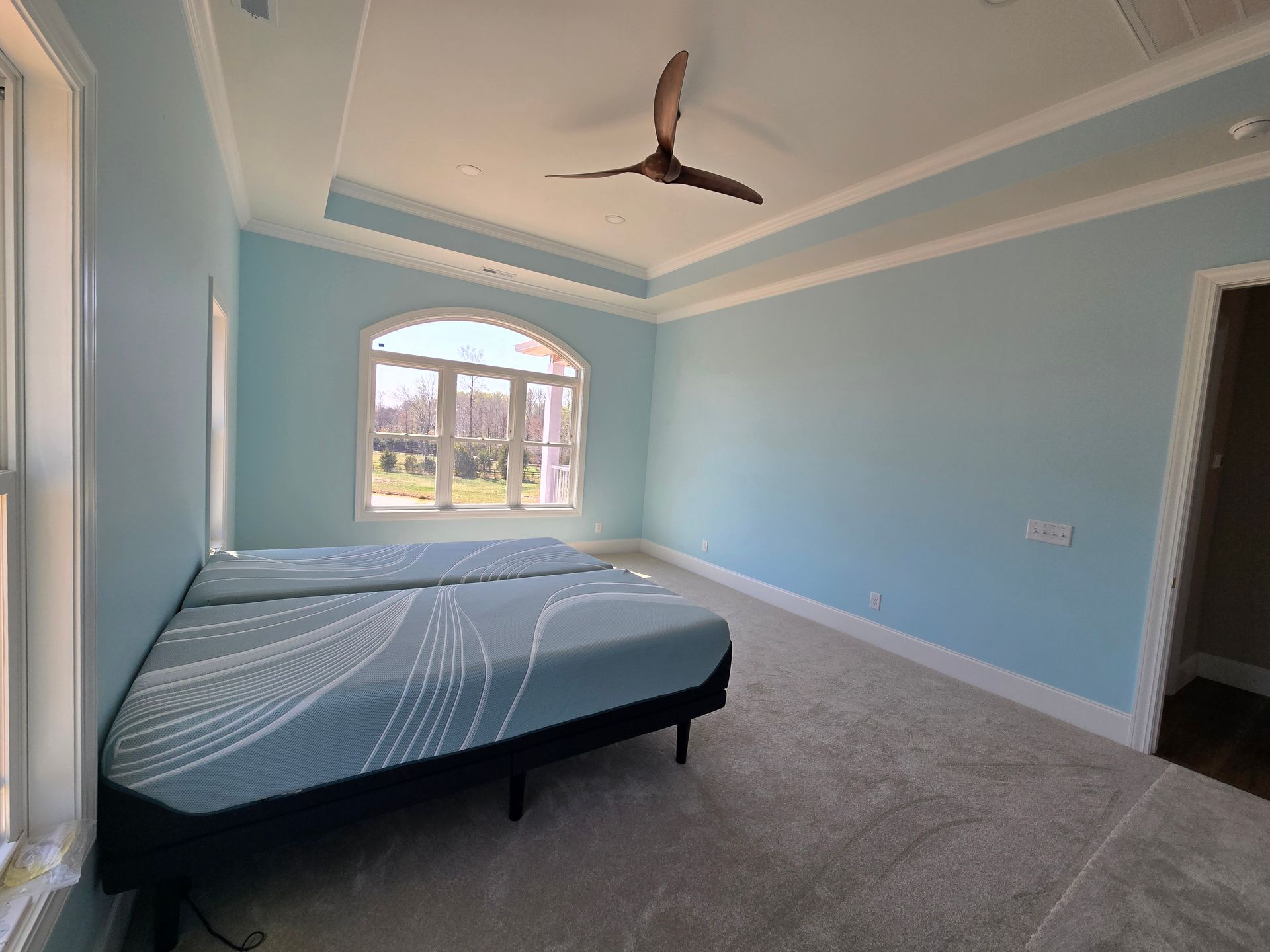 Spacious light blue bedroom with a bed, ceiling fan, large window, and doorway to a bathroom.