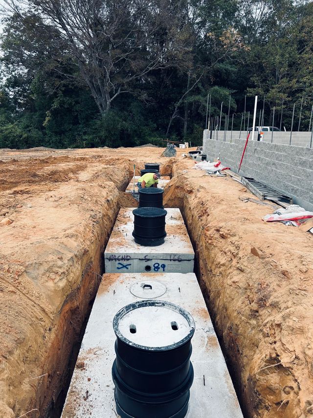 Utility trench with black pipe access caps set in concrete, flanked by dirt excavation at a construction site