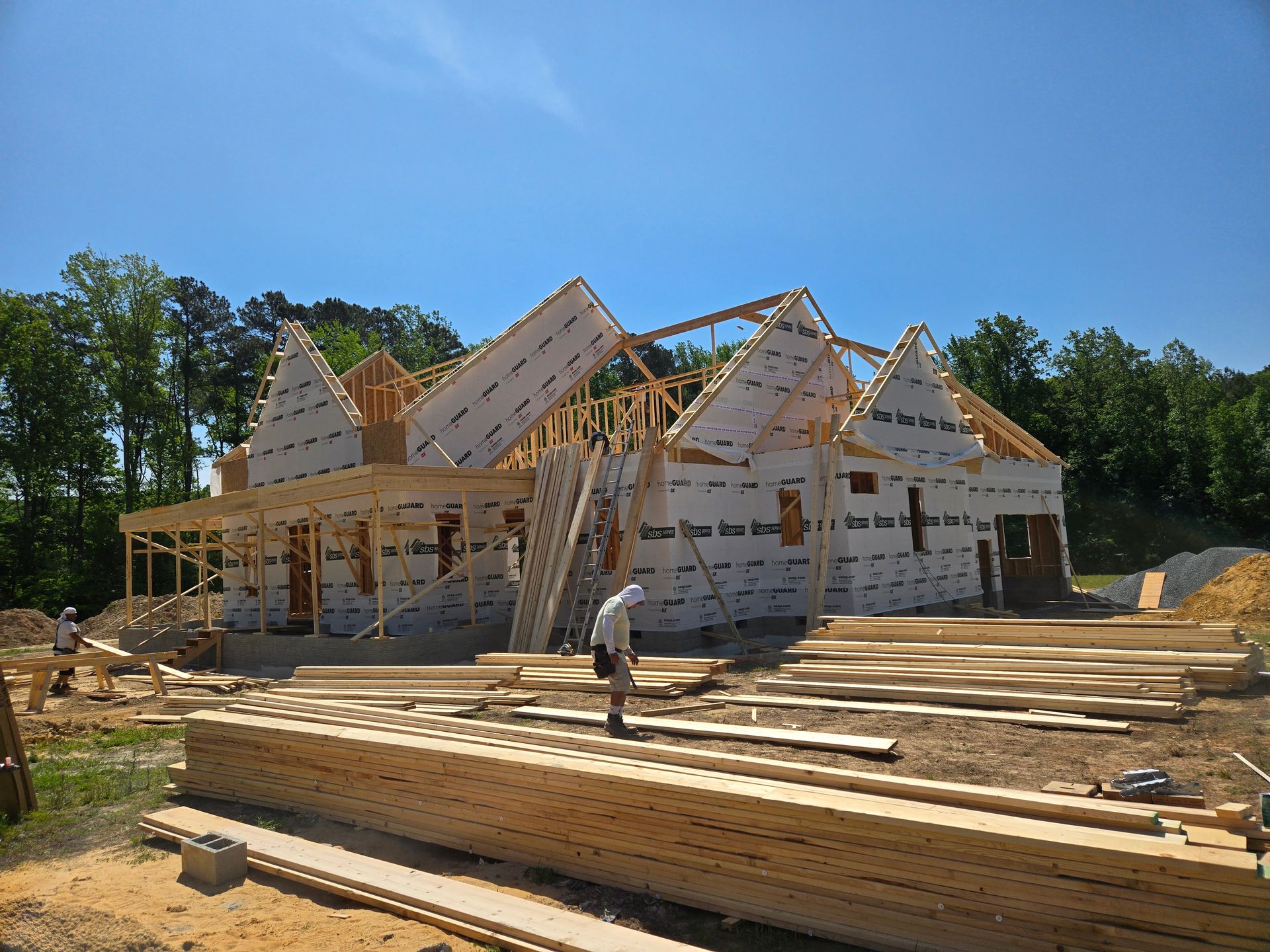 House under construction with wooden framing, stacked lumber, and a worker in front under a clear blue sky