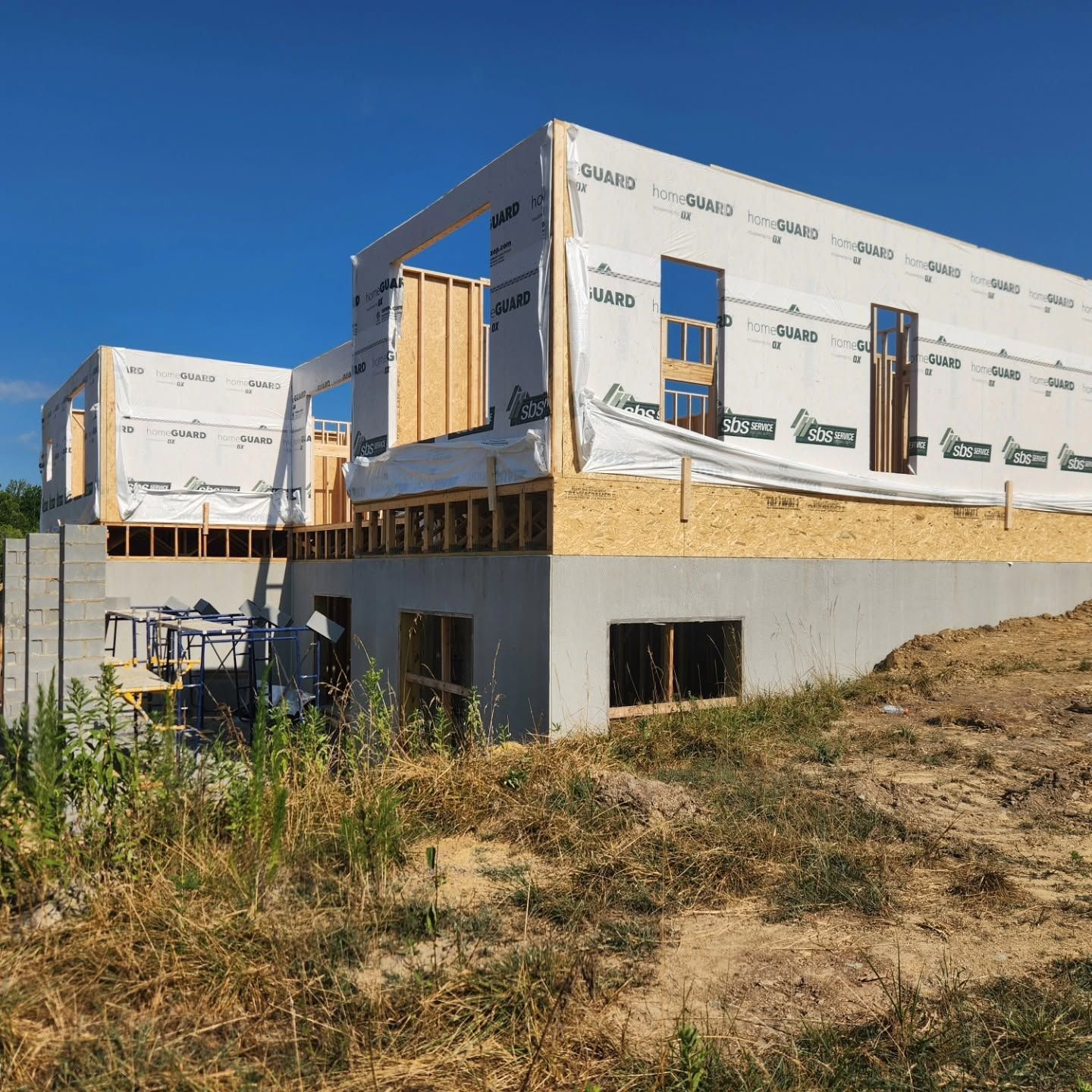 Partially built house with white wrap, wood framing, and overgrown grass under a clear blue sky