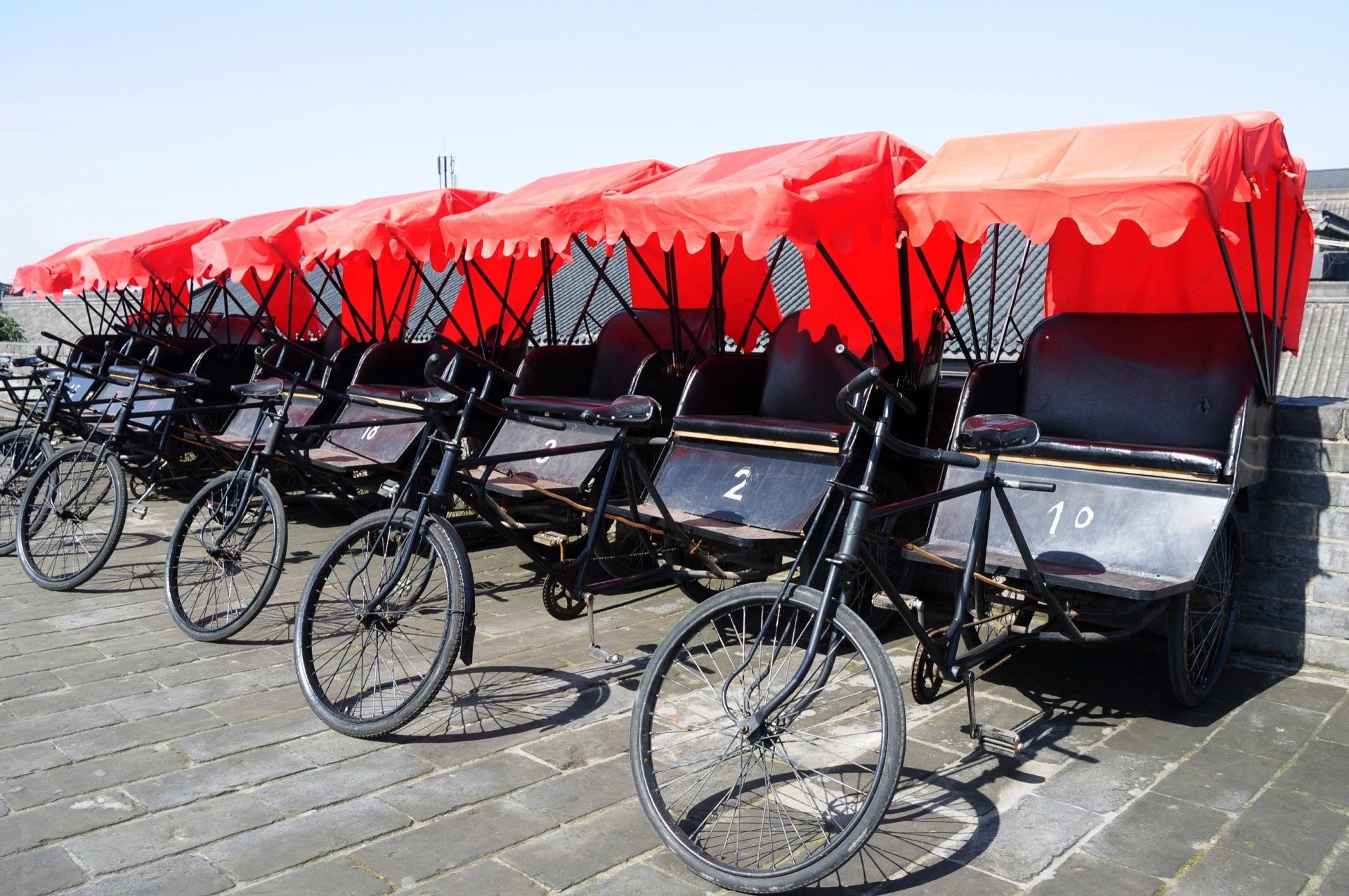 Een rij zwarte fietstaxi's met rode overkappingen staat geparkeerd op een stenen ondergrond.