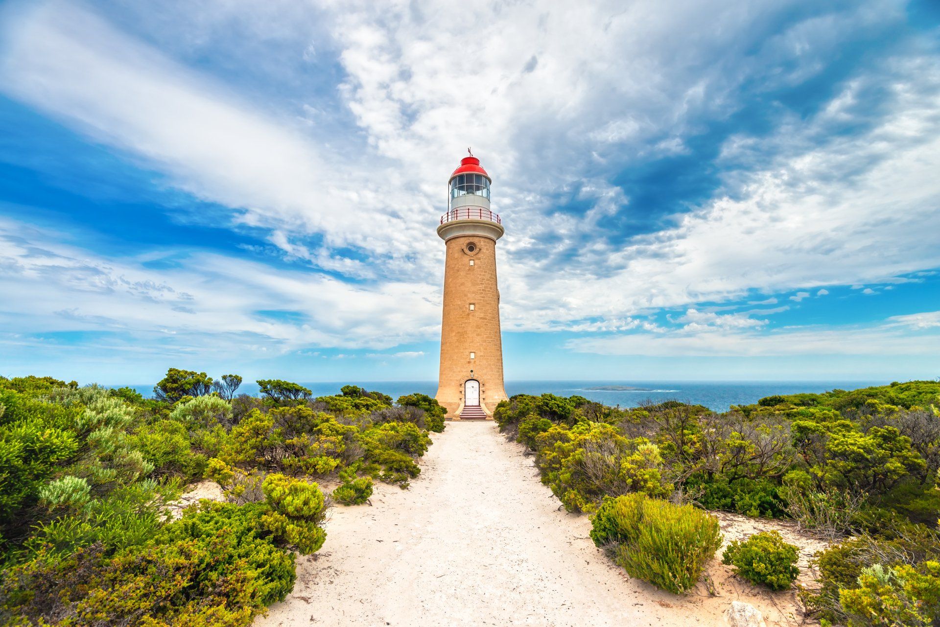 Een vuurtoren op een heuveltop, een pad ernaartoe, een blauwe lucht met wolken en de oceaan op de achtergrond.