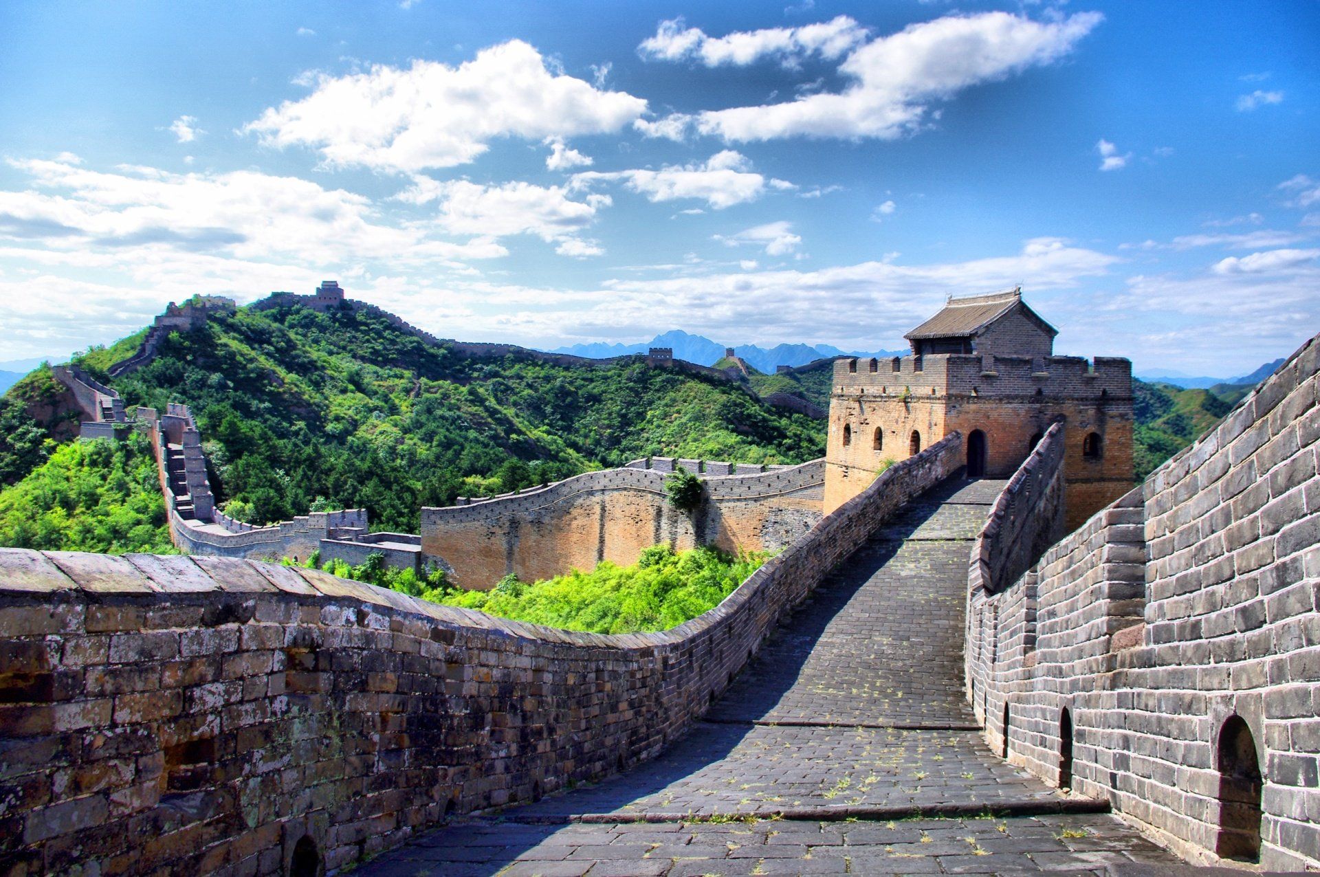 De Grote Muur van China slingert zich door een weelderig, bergachtig landschap onder een blauwe hemel met wolken.