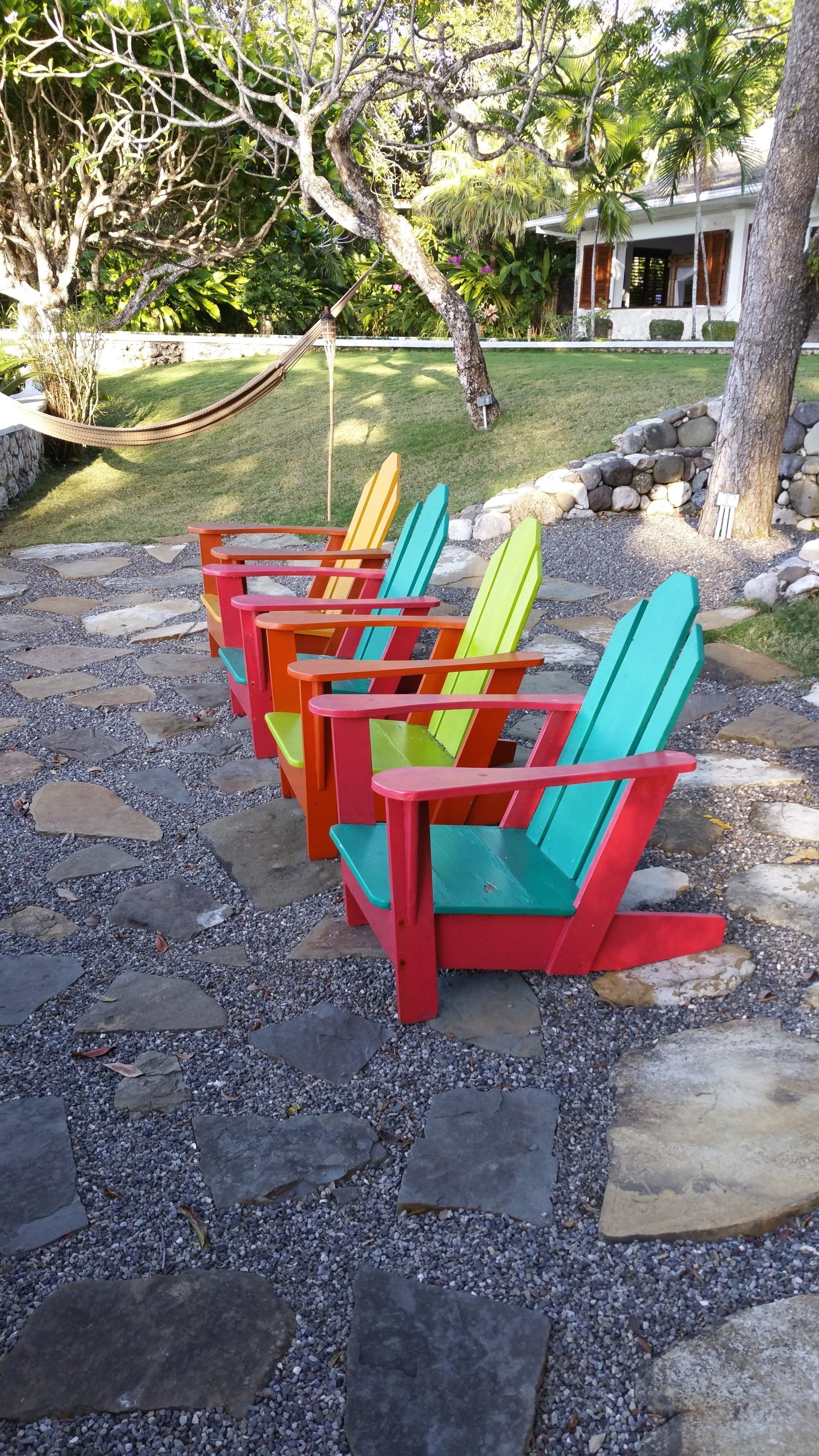 Een rij kleurrijke Adirondack-stoelen op een stenen terras met uitzicht op een grasveld en bomen.