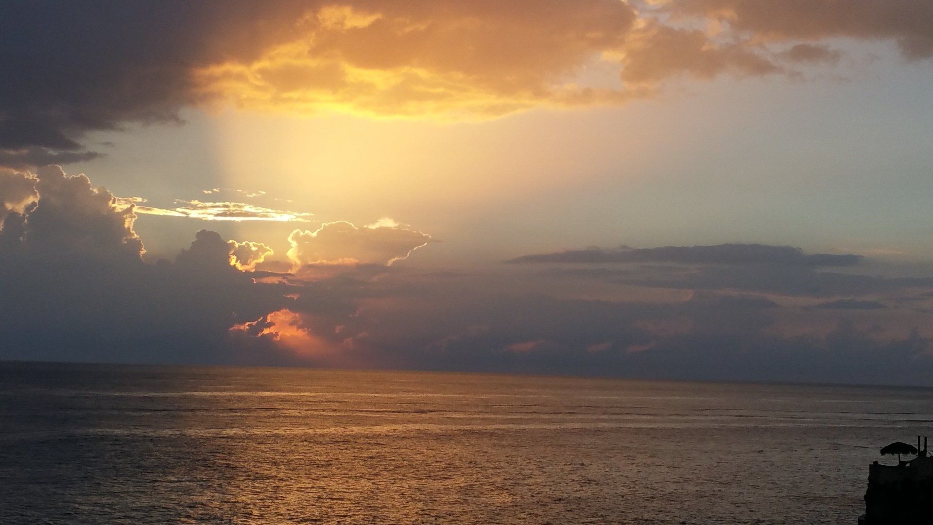 Zonsondergang boven de oceaan met zonnestralen die door de wolken breken; goud- en oranjetinten.