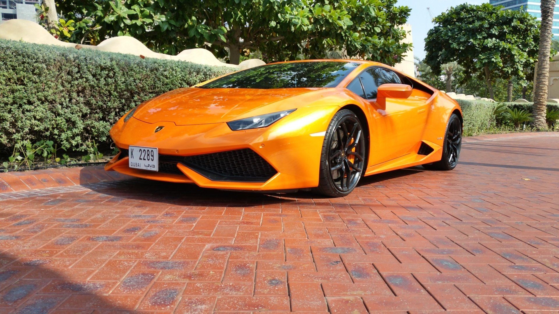 Een oranje Lamborghini sportwagen staat geparkeerd op een met bakstenen bestraat terrein, vlakbij een groenstrook.