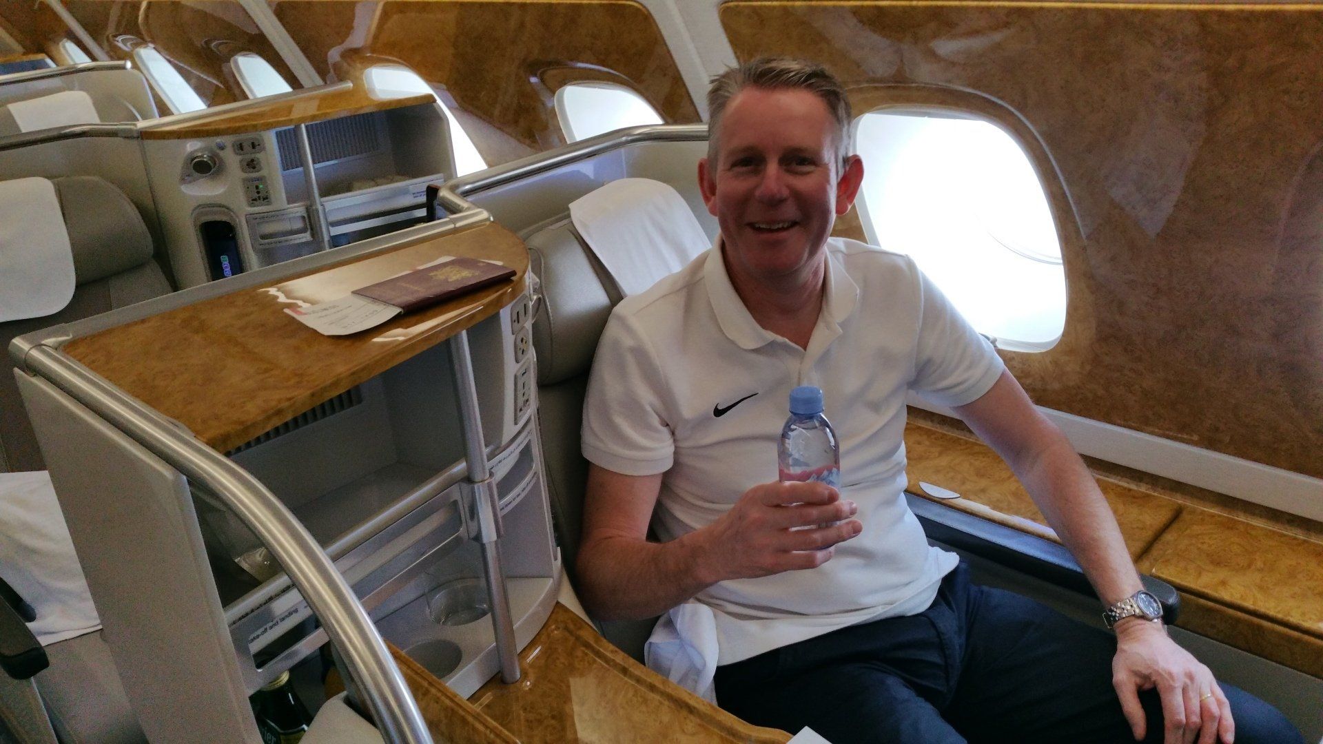 Een man glimlacht en houdt een fles water vast in een privéjet. Luxe cabine met hout en leer.