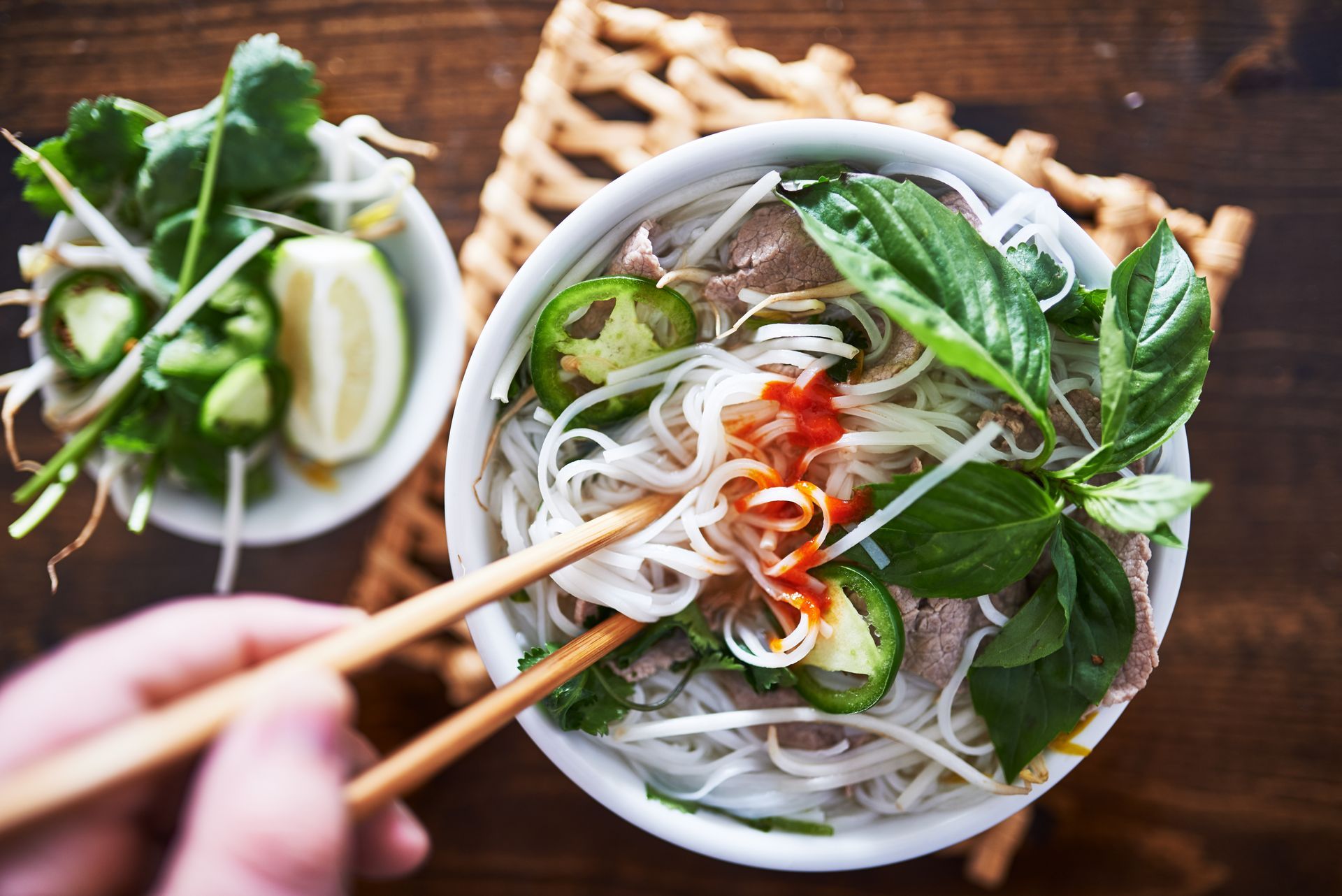 Een kom pho met eetstokjes, kruiden en toppings, geserveerd op een houten tafel.