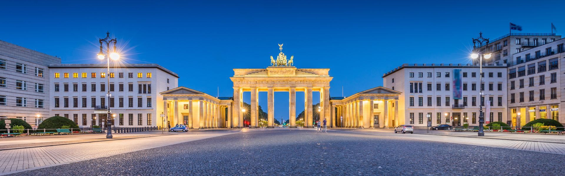 De Brandenburger Tor in Berlijn, verlicht in de schemering. Gebouwen aan weerszijden van de poort met een geplaveid plein.