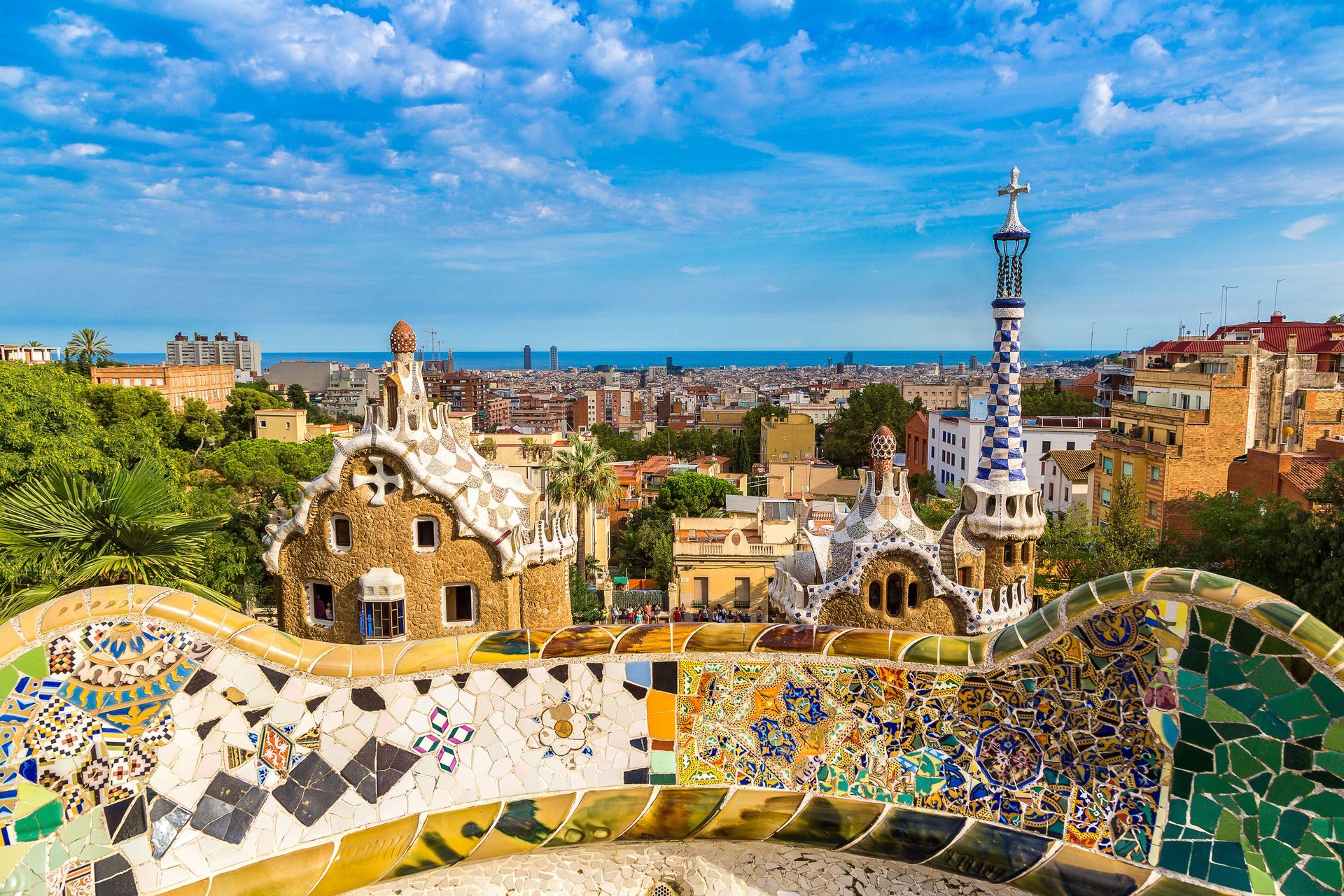 Park Güell in Barcelona, ​​Spanje, kenmerkt zich door kleurrijke mozaïekbanken en unieke architectonische structuren tegen een stadsgezicht en een blauwe hemel.