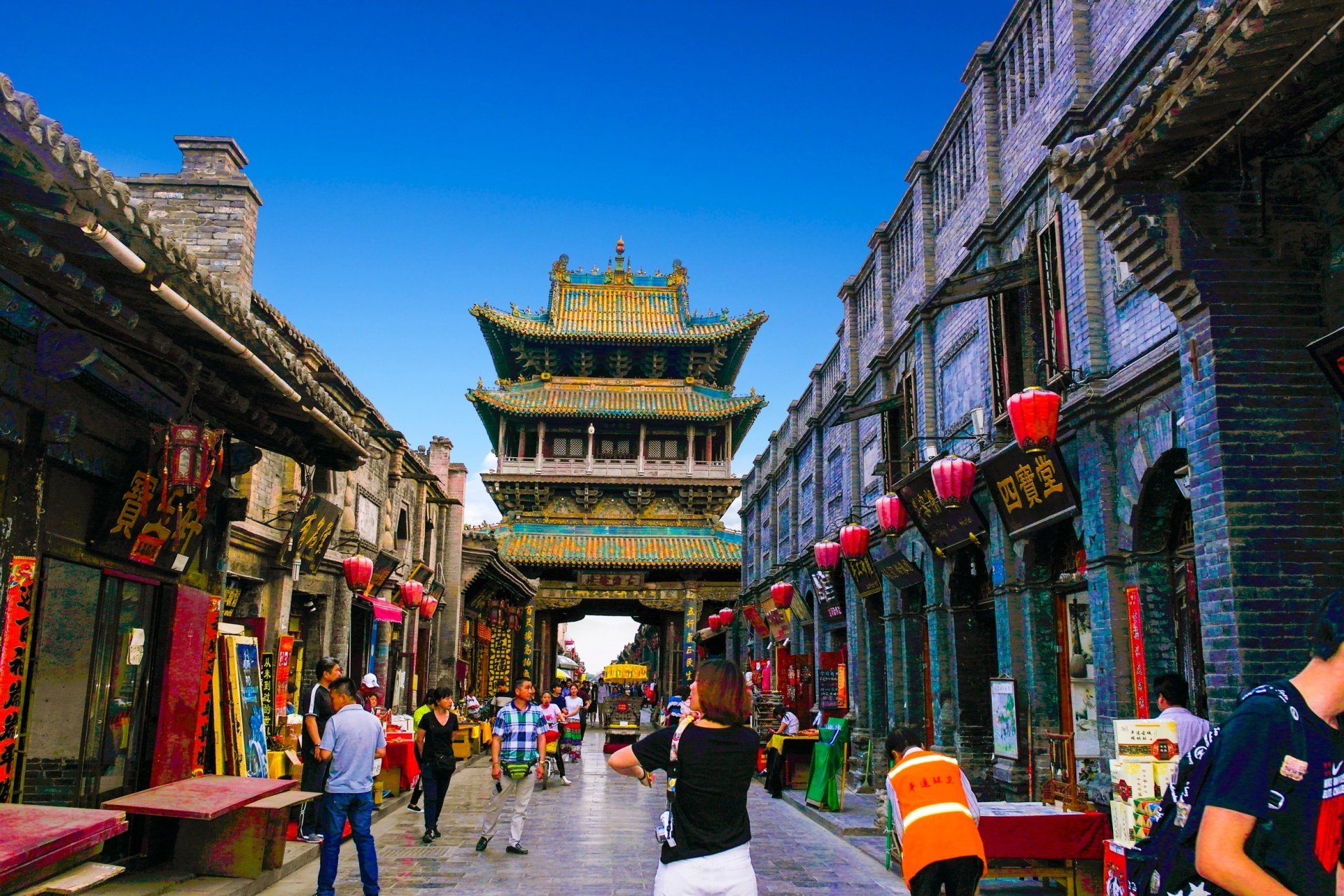 Straatbeeld in China met een grote, sierlijke poort en winkels, mensen die onder een blauwe hemel lopen.