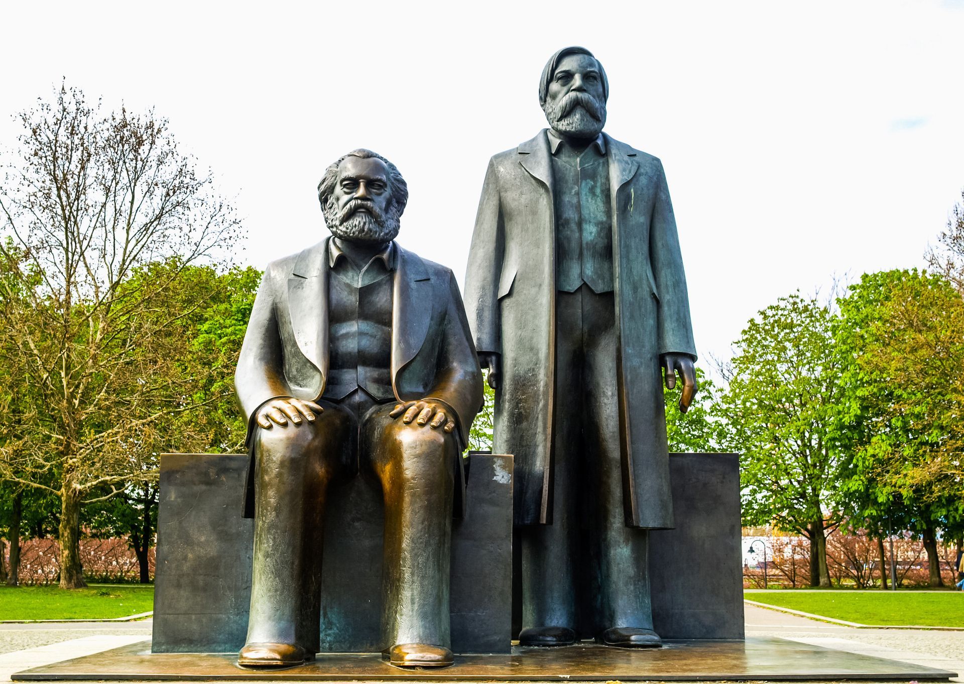 Bronzen beeld van Karl Marx zittend en Friedrich Engels staand, in een parkomgeving.