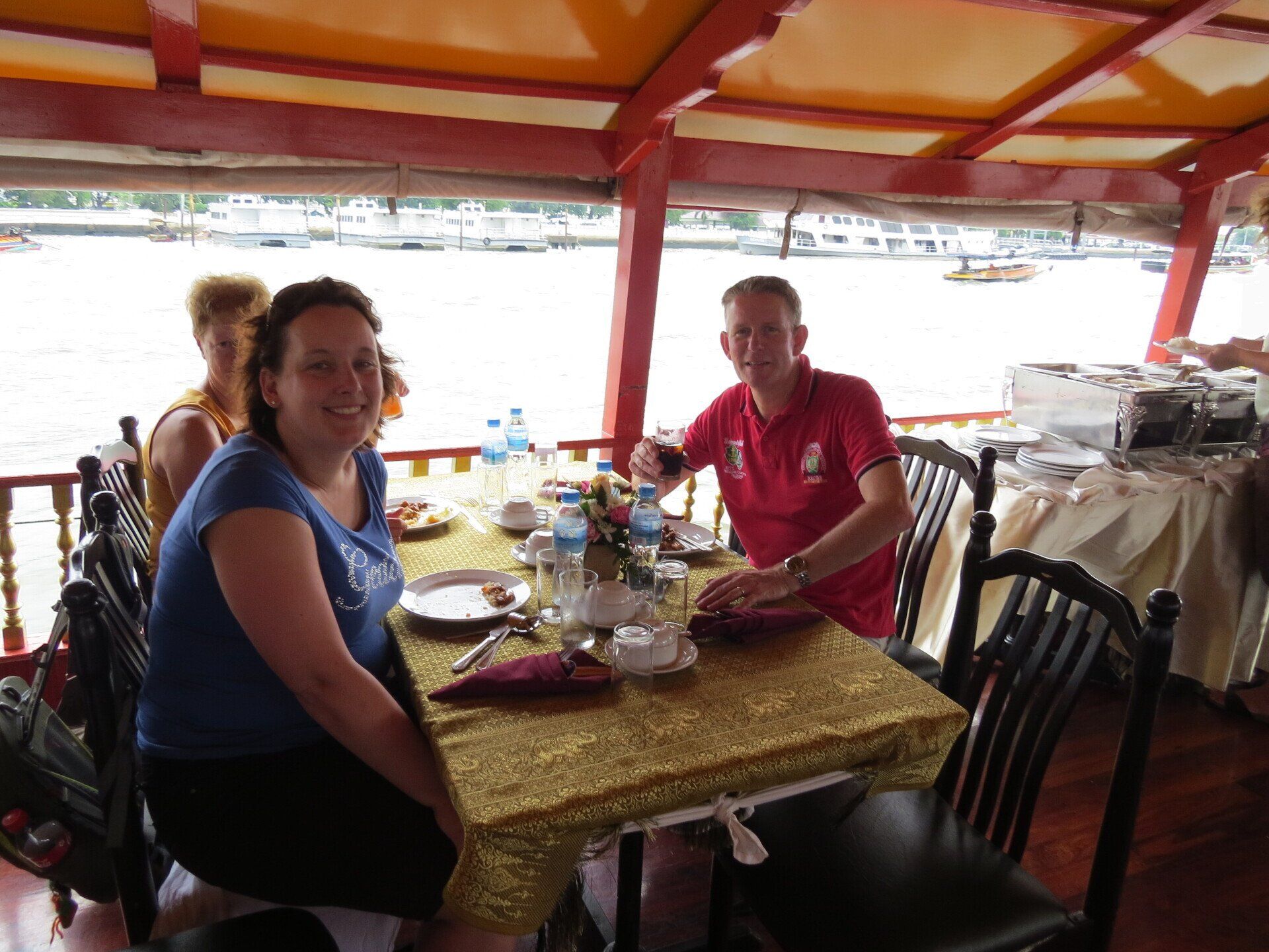 Mensen zitten aan een tafel op een boot te eten. Vrouw in blauw, man in rood shirt. Buffet op de achtergrond.