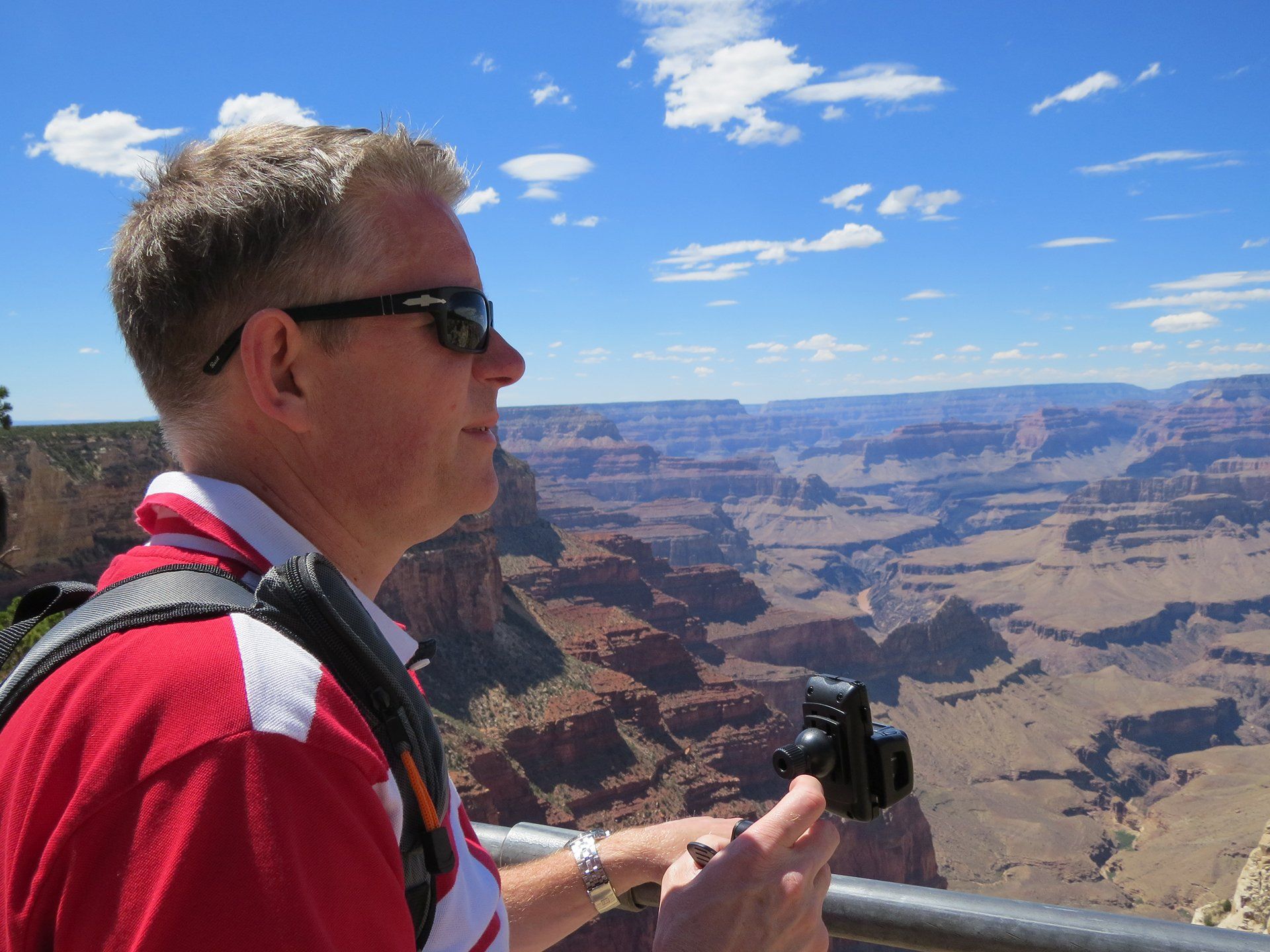 Een man in een rood shirt en met een zonnebril kijkt naar de Grand Canyon en houdt een camera vast. Blauwe lucht, zonnige dag.