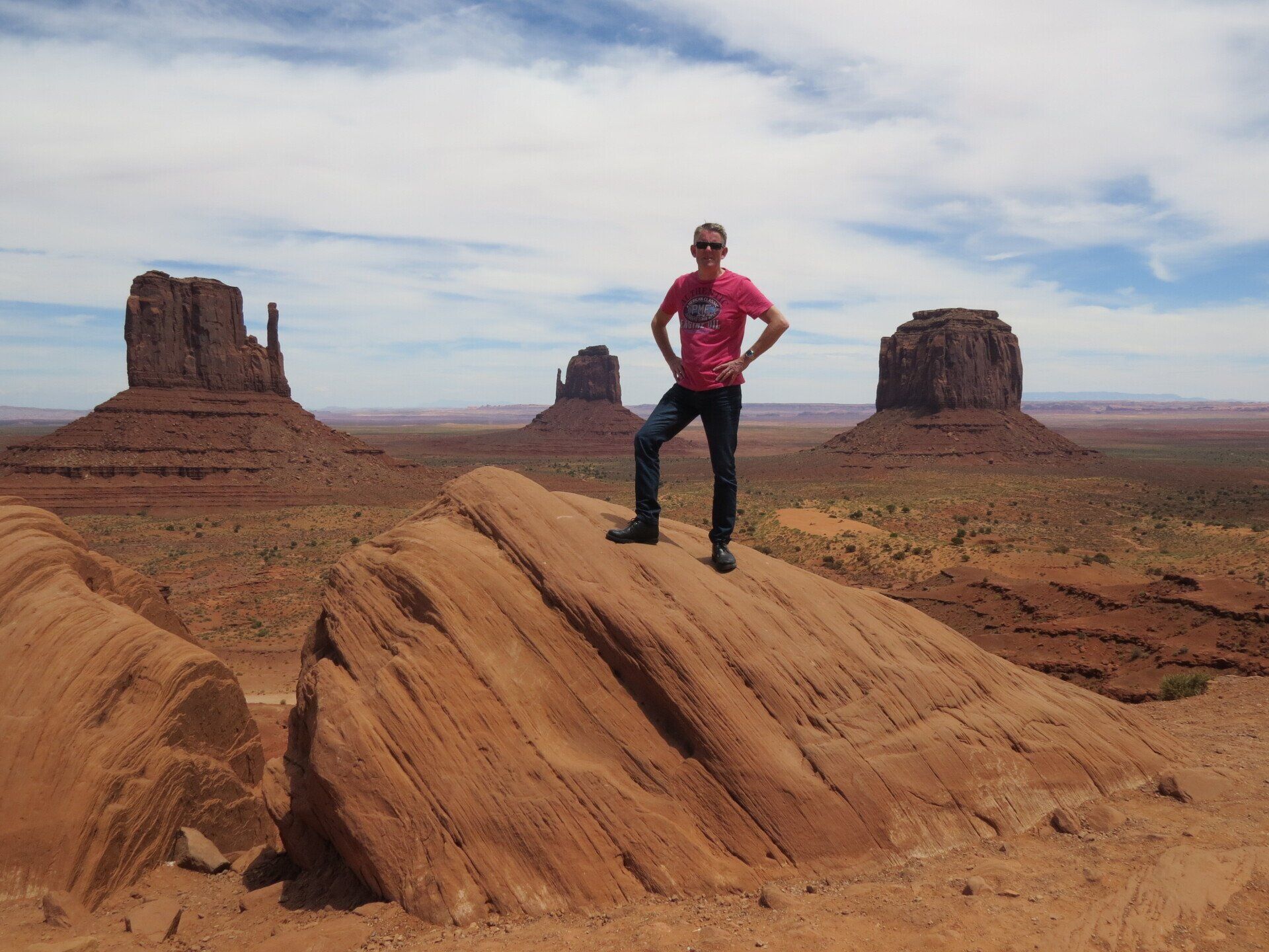 Een man staat op een rotsformatie in Monument Valley, met zijn handen in zijn zij, rotsformaties op de achtergrond en een blauwe lucht.