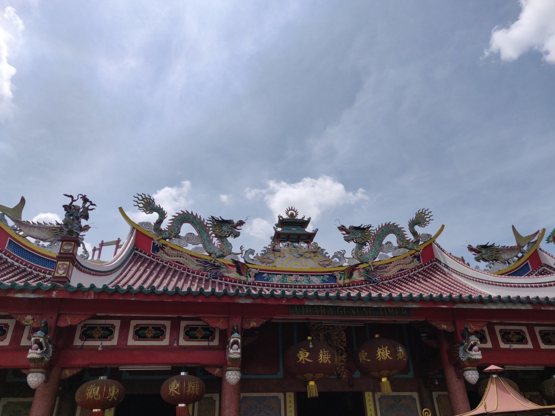 Rood-wit Chinees tempeldak met drakensculpturen en lantaarns, tegen een bewolkte hemel.