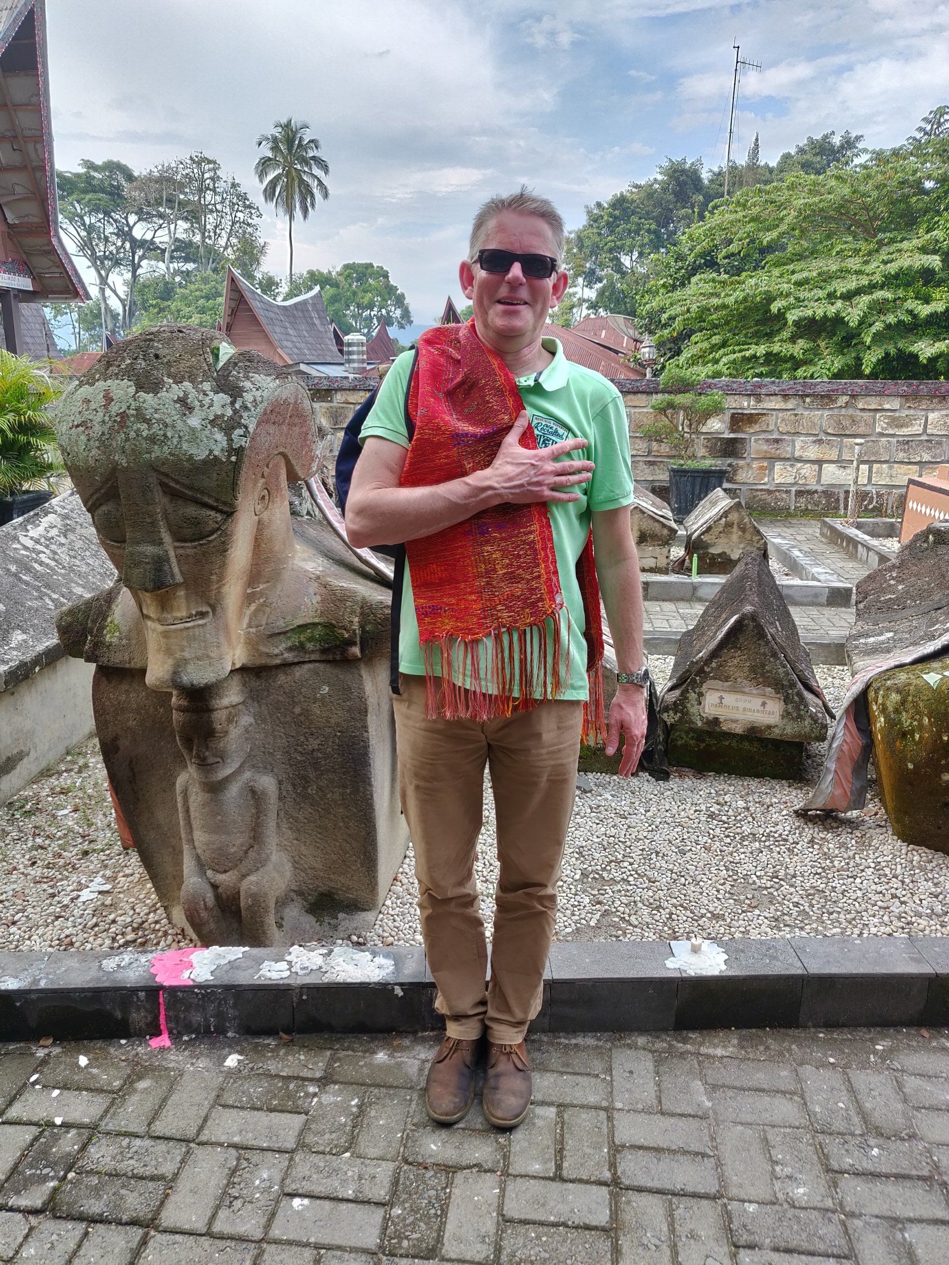 Een man in een groen shirt en met een sjaal om staat voor oude stenen bouwwerken en houdt zijn hand op zijn borst.
