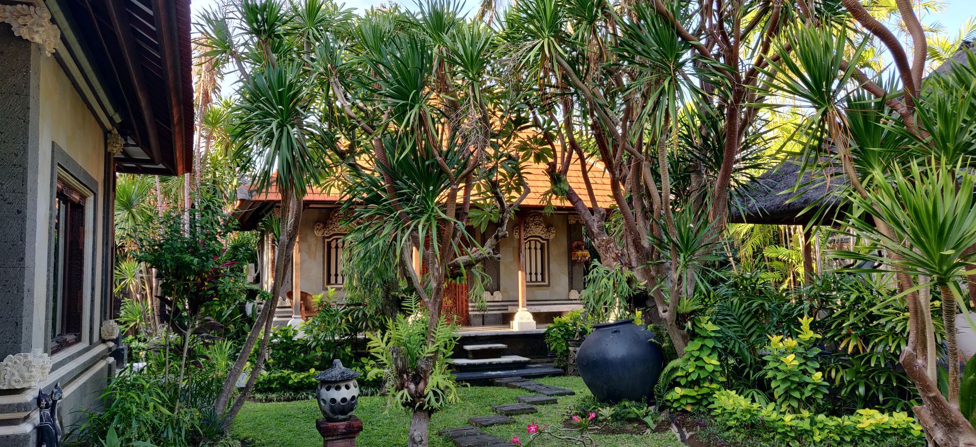 Tuin met tropische planten, traditionele gebouwen, een stenen pad en een decoratieve pot.