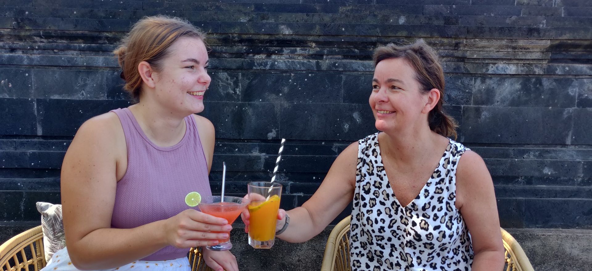Twee vrouwen klinken met een glimlach op hun gezicht hun cocktailglazen terwijl ze aan een tafel zitten. Op de achtergrond is een donkere muur te zien.