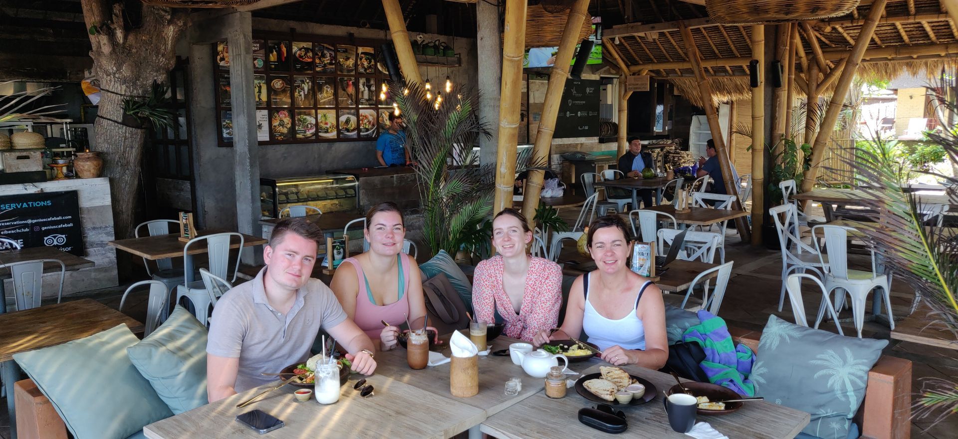 Vier mensen zitten aan een tafel in een café en genieten van eten en drinken. De setting is buiten en heeft een rustieke uitstraling.