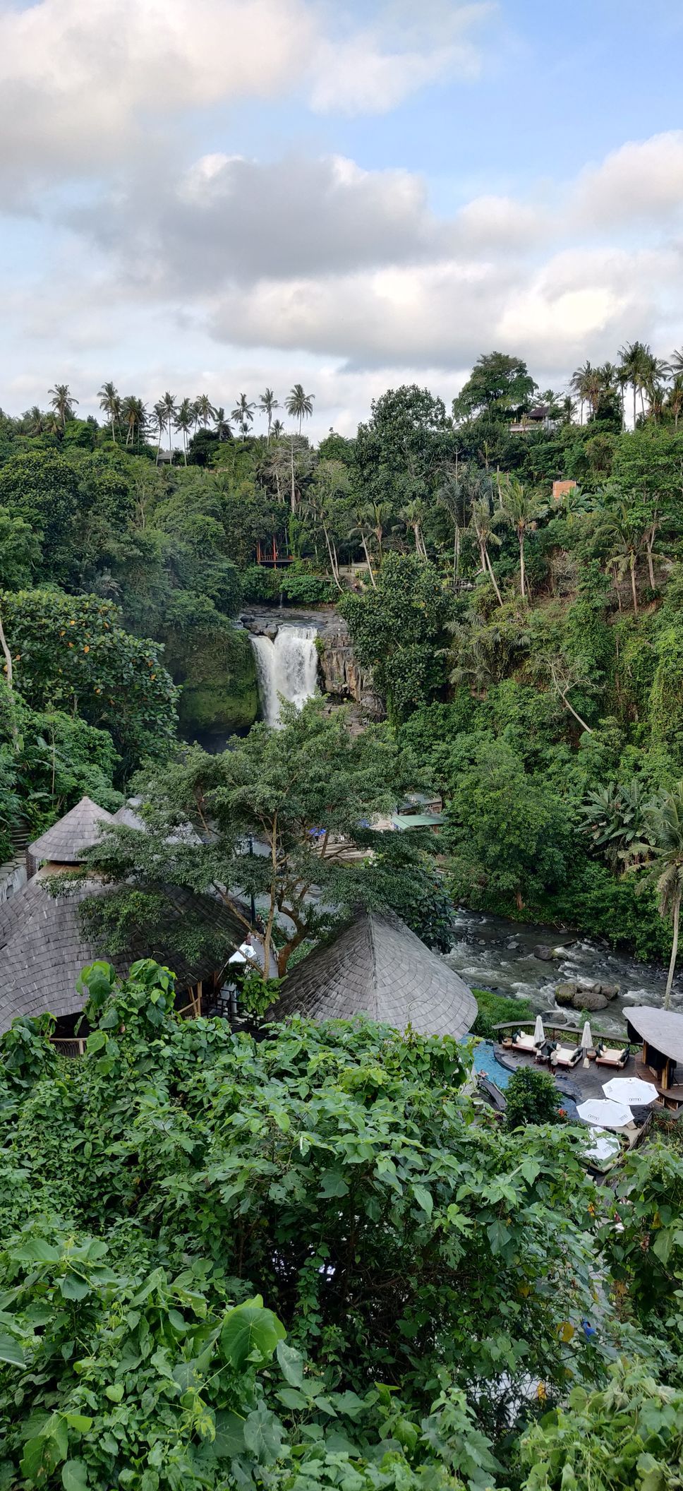 Uitzicht op een waterval die door weelderig groen naar beneden stort, met hutten zichtbaar op de voorgrond.