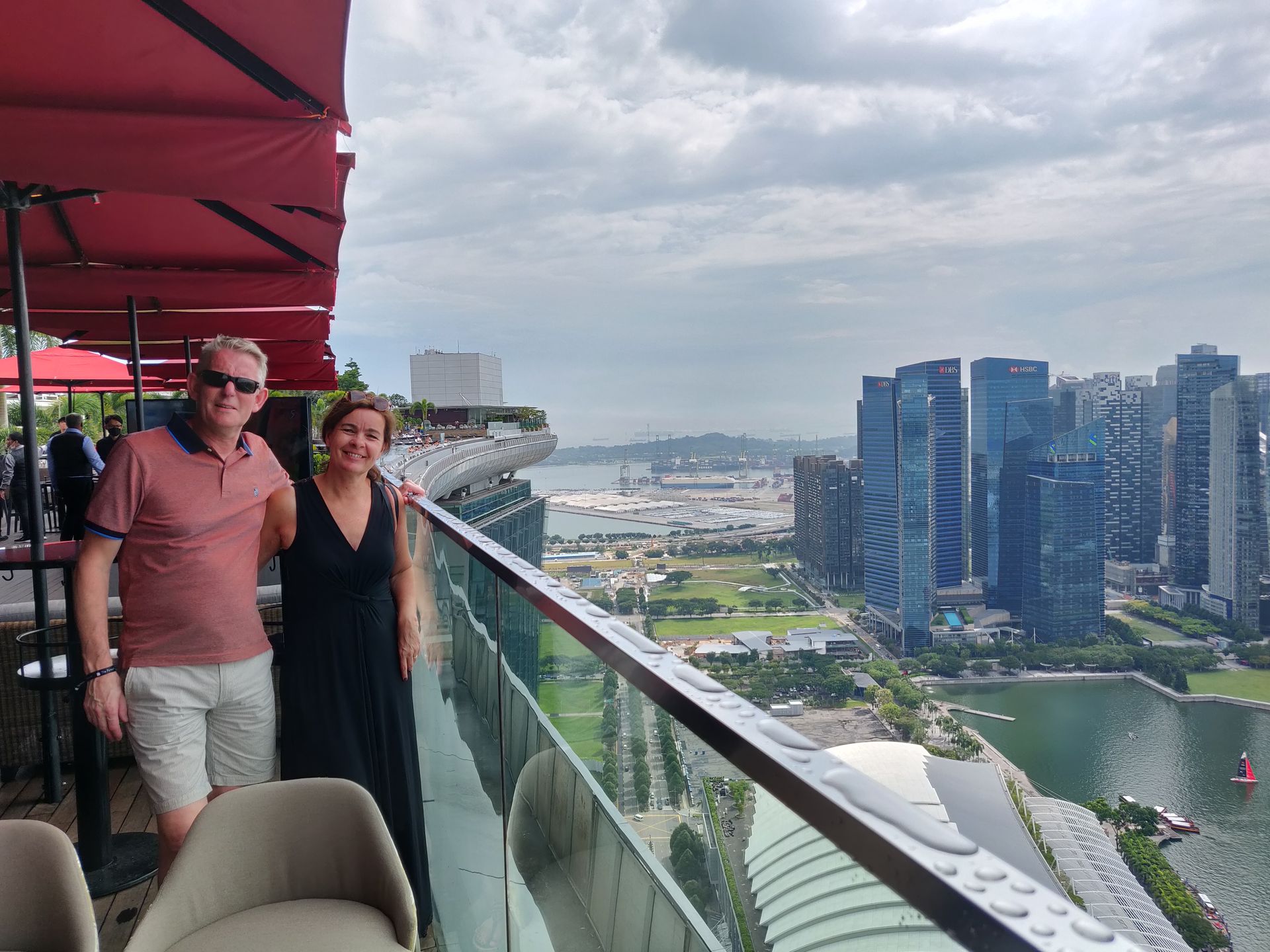 Een stel op een balkon van een hoog gebouw, met uitzicht op de skyline van de stad en de haven op een bewolkte dag.