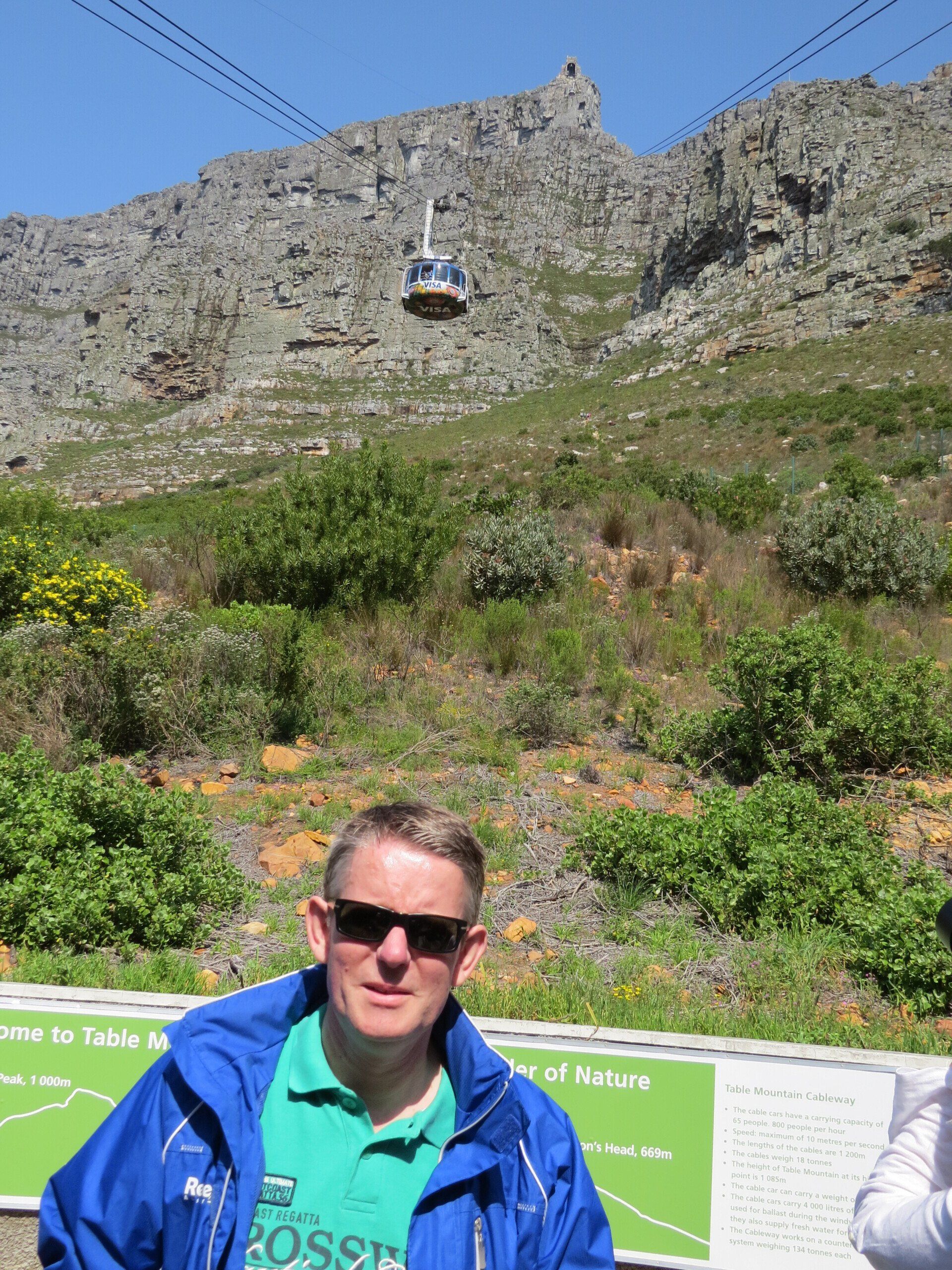 Man met zonnebril en blauwe jas, kabelbaan naar de Tafelberg op de achtergrond.