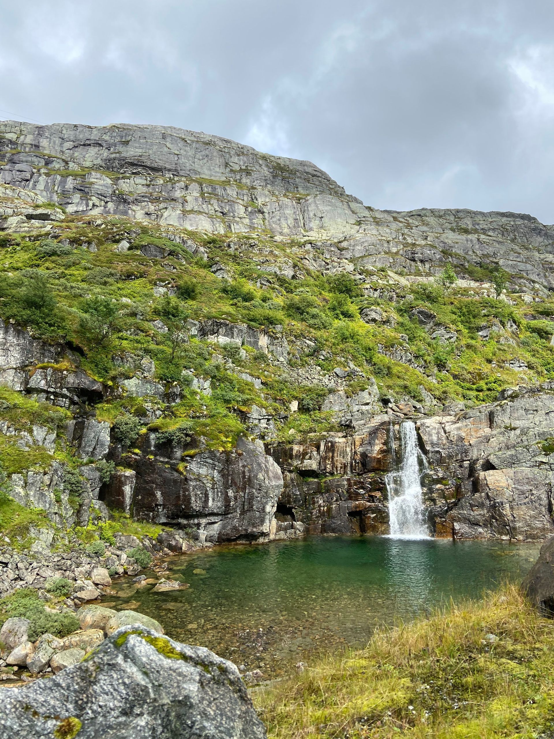Een waterval die neerstort in een turquoise poel, omgeven door rotsachtige kliffen en groene vegetatie onder een bewolkte hemel.