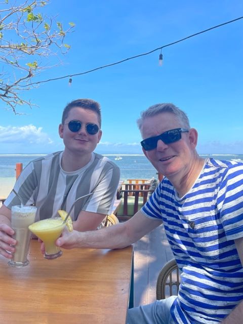 Twee mensen zitten aan een tafeltje buiten, elk met een drankje in de hand. Op de achtergrond zie je de oceaan en een blauwe lucht.