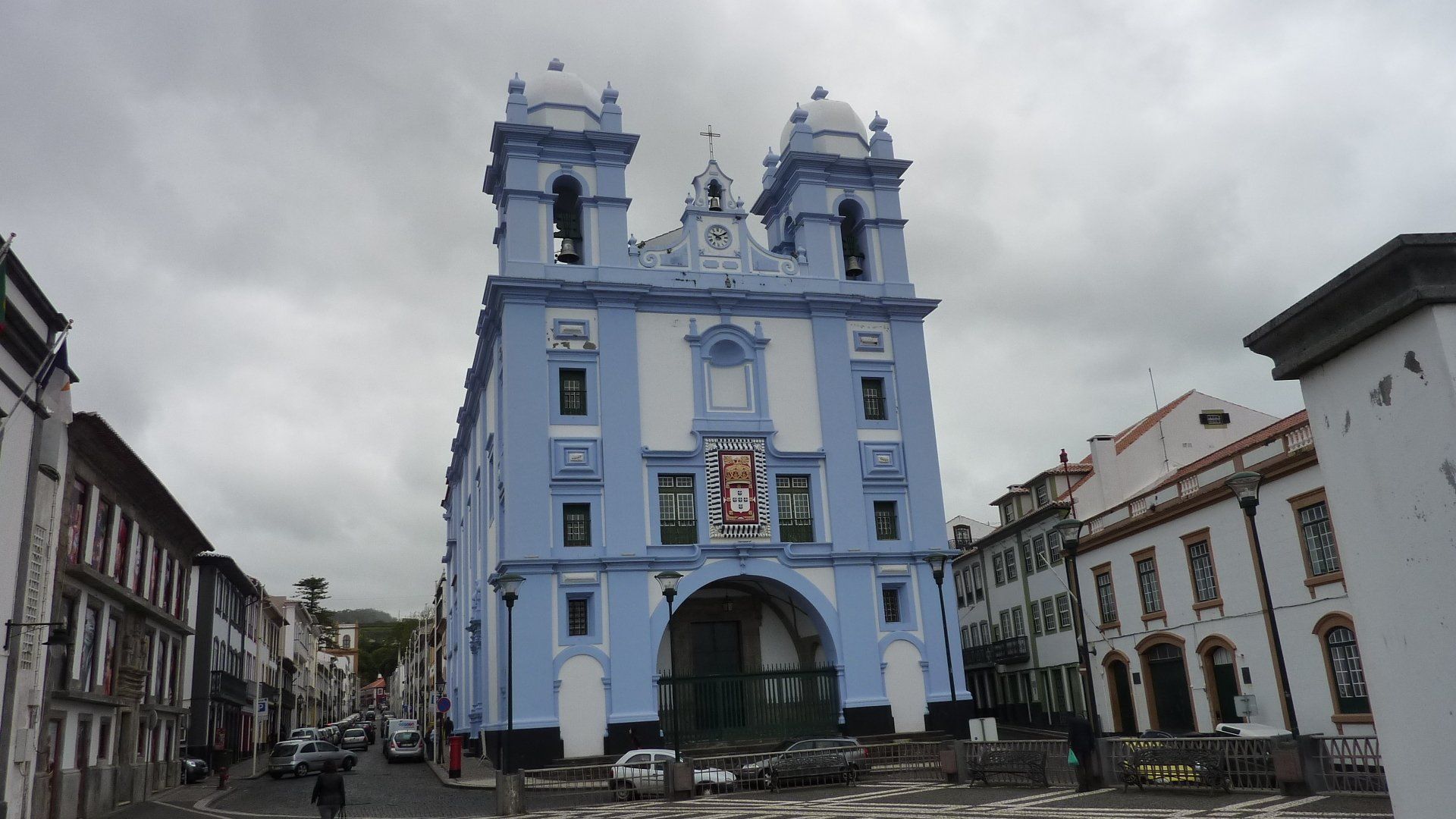 Een blauw-witte kerk met twee klokkentorens op een Europees stadsplein. Bewolkte lucht erboven.