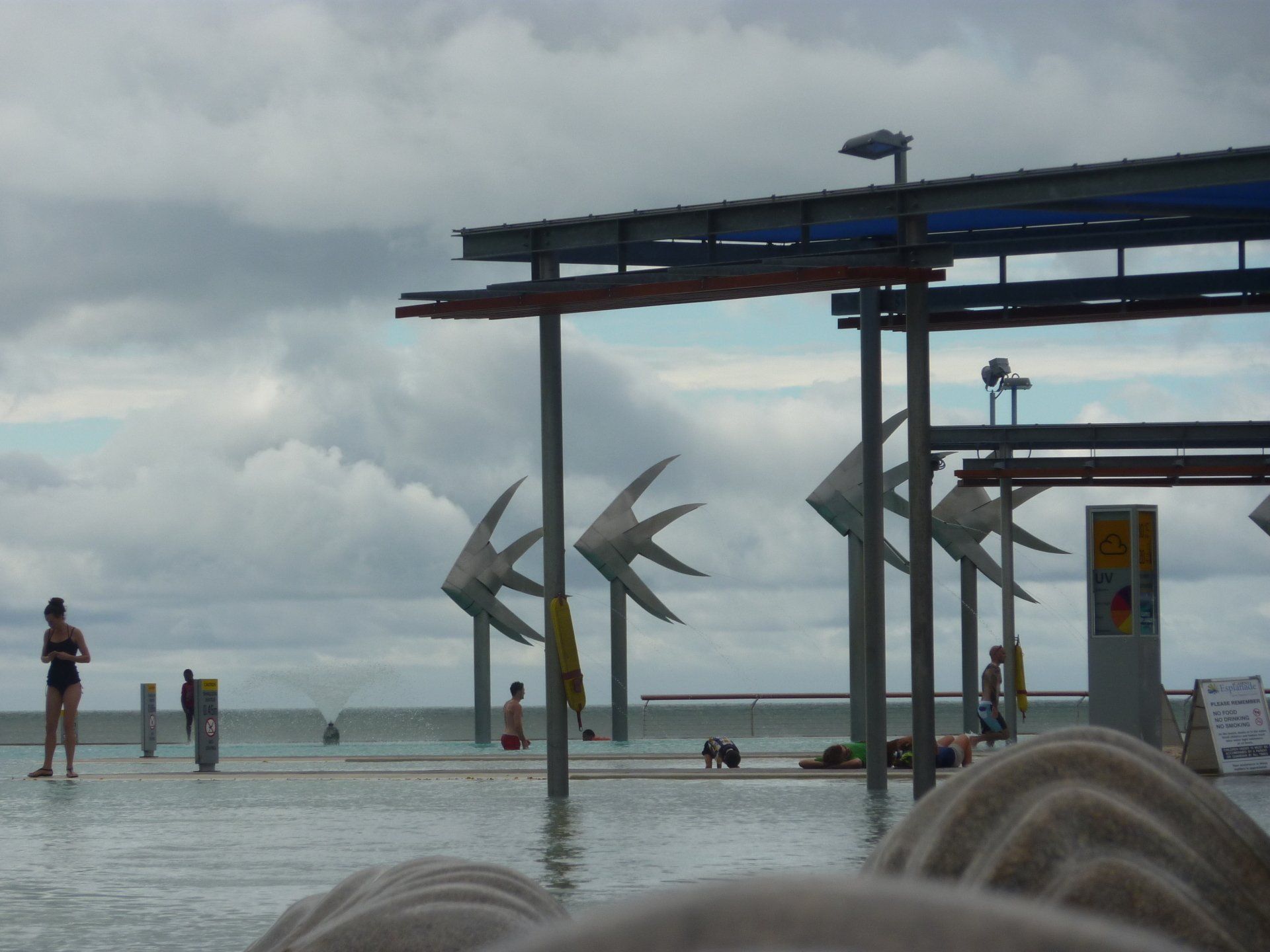 Openbaar zwembad met vissculpturen en een bewolkte hemel. Mensen zwemmen en ontspannen.