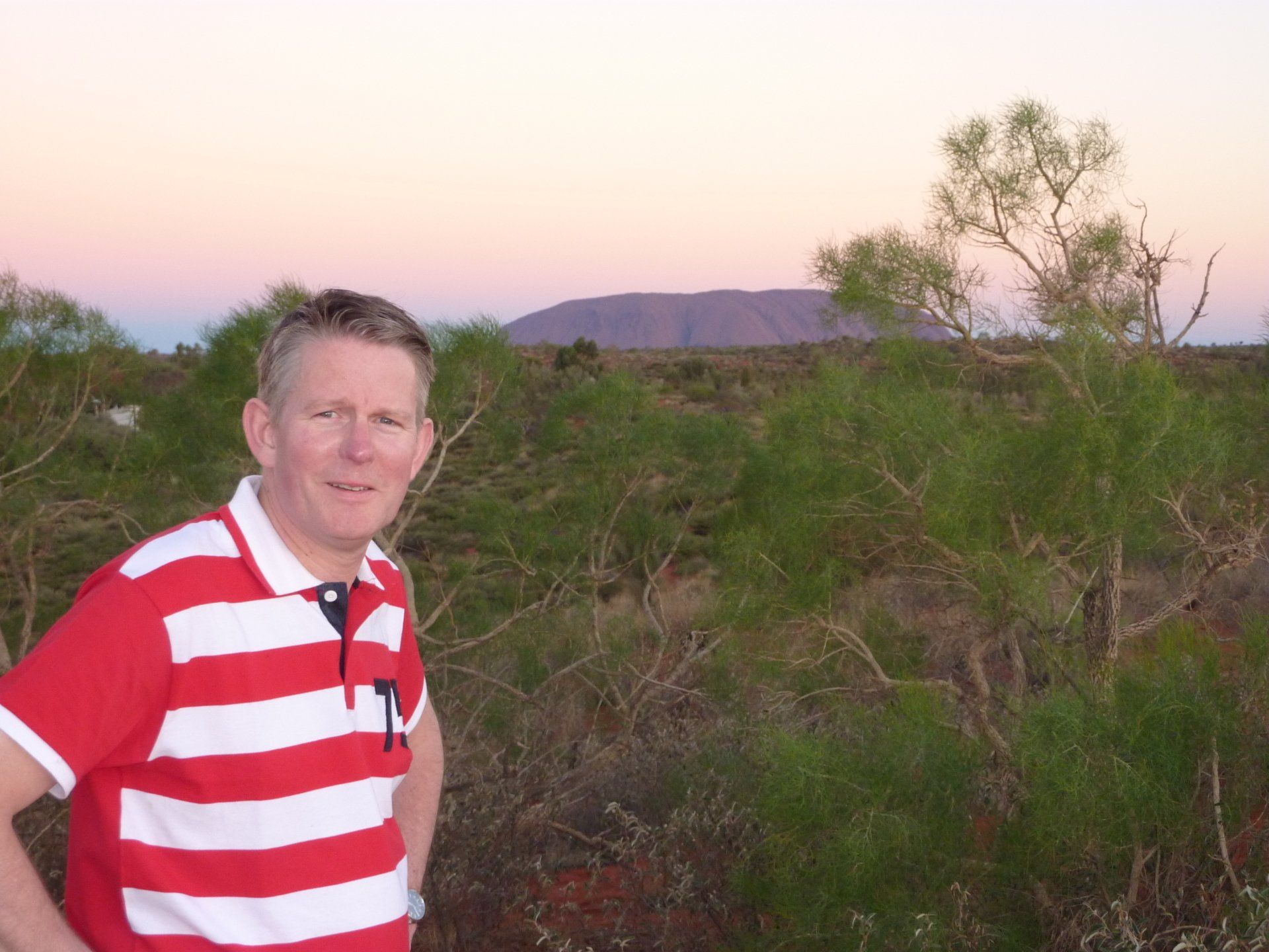 Een man in een rood-wit gestreept shirt staat bij struiken, met een berg in de verte. De kleuren van de zonsondergang sieren de lucht.