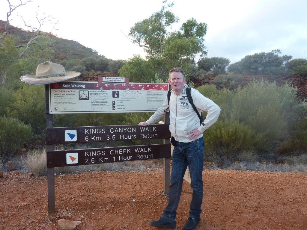 Een man staat bij een bord met wandelroute-informatie in Kings Canyon. Op het bord staan ​​de loopafstanden aangegeven. De man draagt ​​een beige hoed, die zich in de outback bevindt.