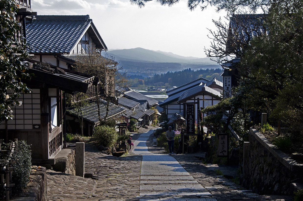 Een stenen straat met traditionele Japanse gebouwen; in de verte zijn bergen te zien.