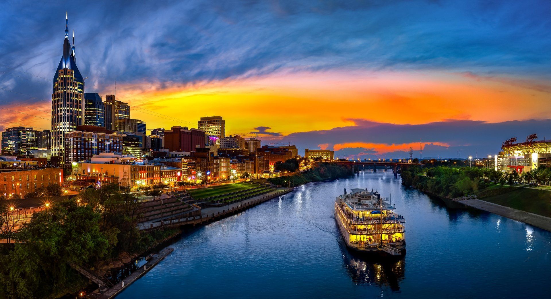 De skyline van Nashville bij zonsondergang, met een rivier en een verlichte boot. Een oranje en blauwe lucht boven de stad.