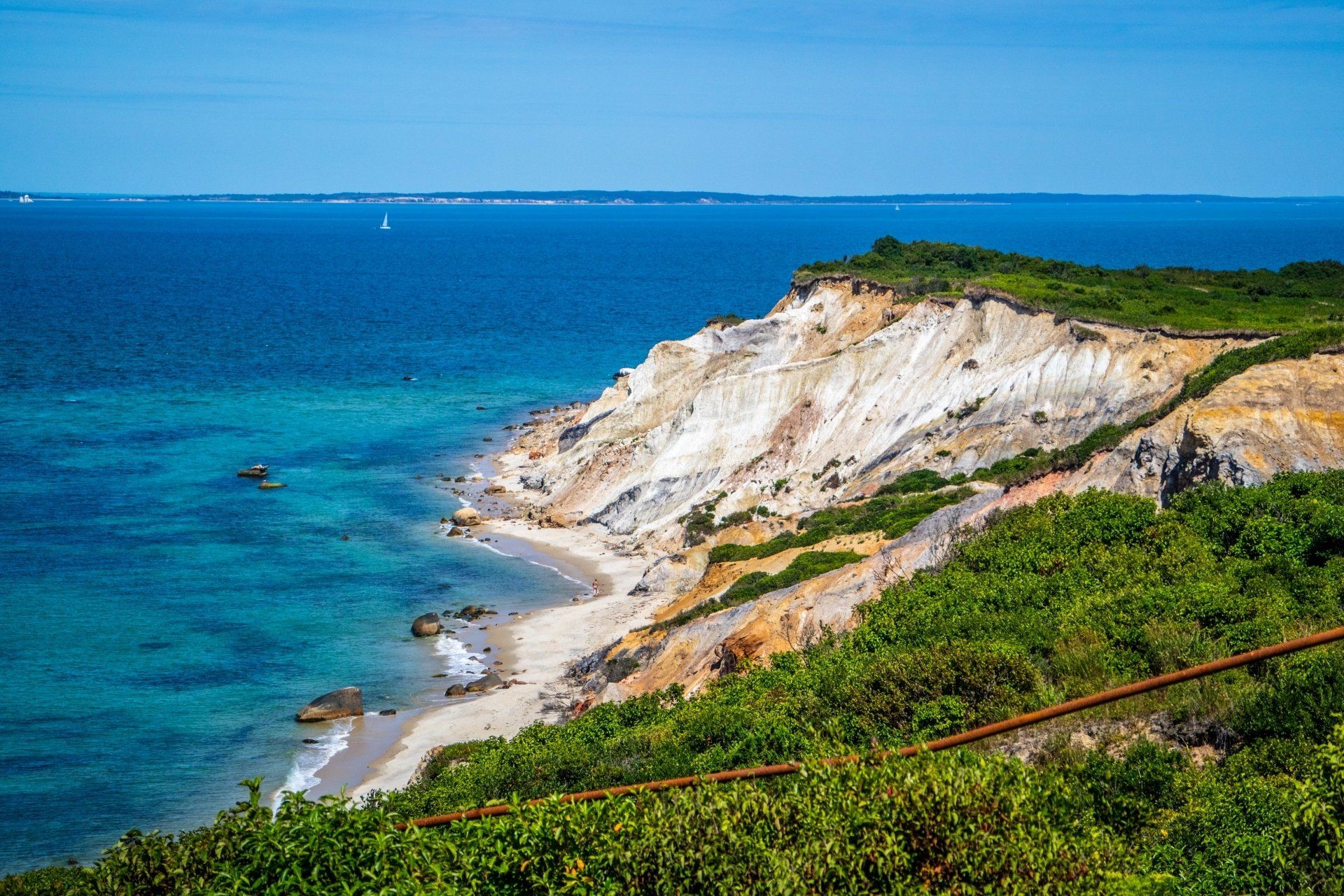 Kliffen aan de kust met uitzicht op een turquoise oceaan onder een blauwe hemel; zandstrand en groene vegetatie zijn zichtbaar.