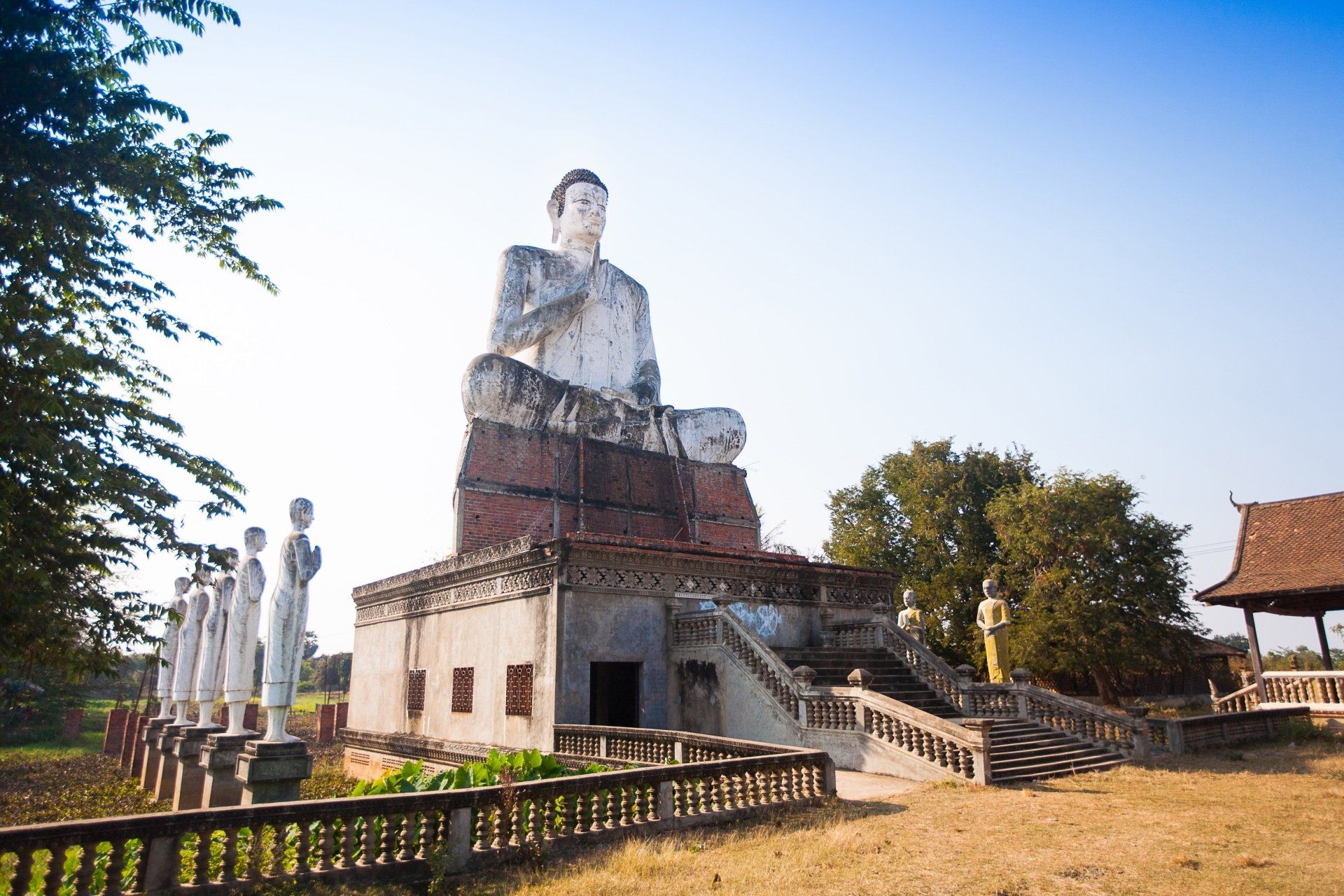 Vervallen tempel met een groot wit Boeddhabeeld en beelden van andere figuren. Droog veld, blauwe lucht.