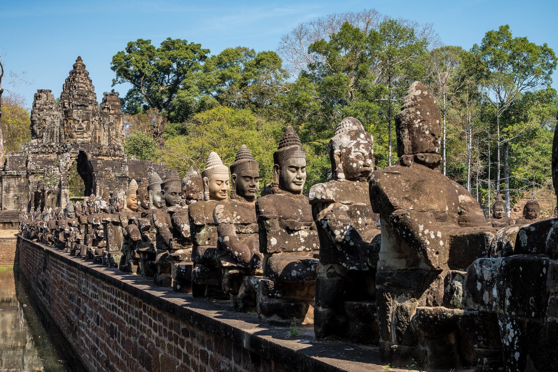 Stenen beelden staan ​​langs een brug die naar een oude tempel in Cambodja leidt; blauwe lucht, bomen op de achtergrond.