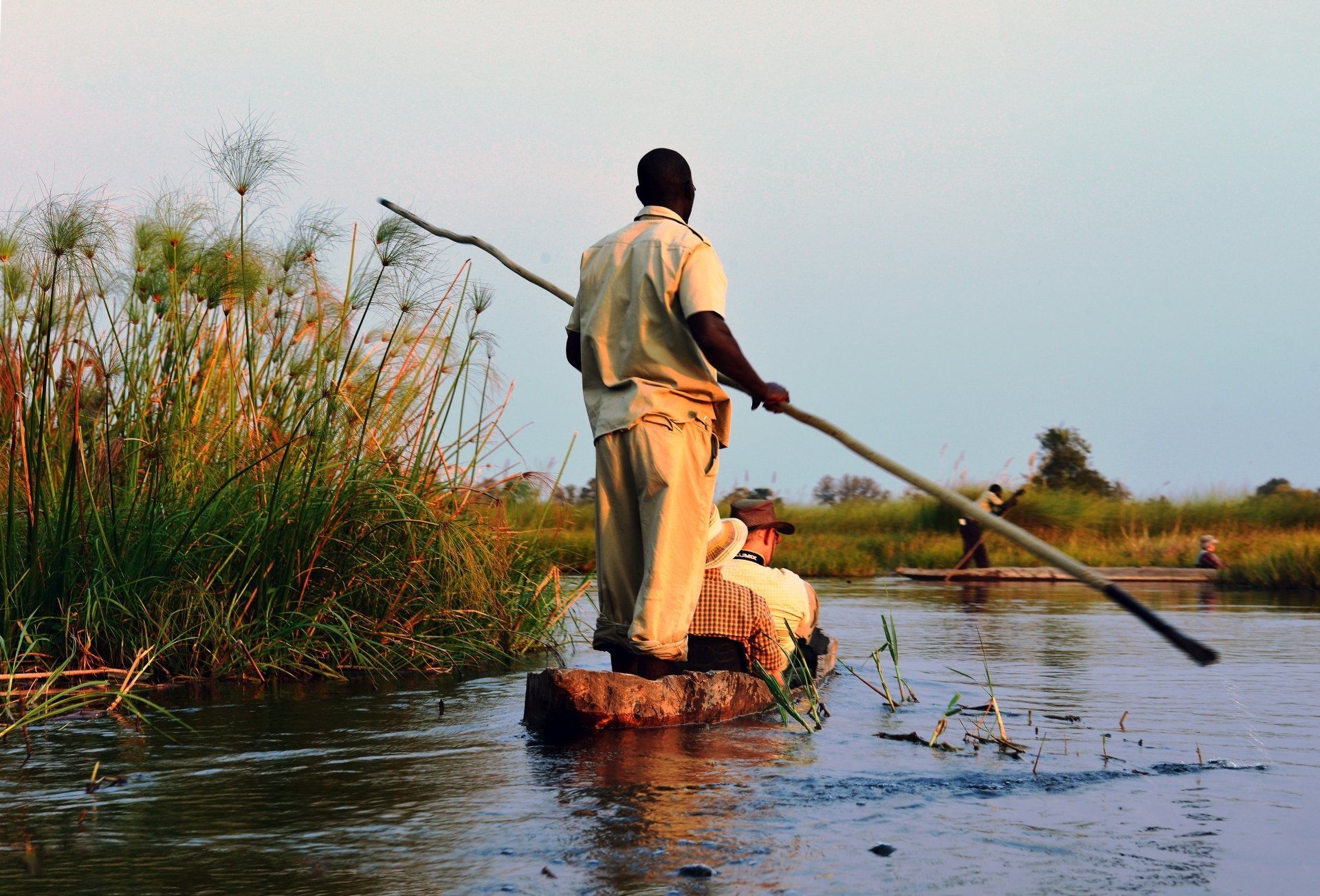 Een man duwt een mokoro-boot door het water, in de verte is een andere boot te zien, links staat hoog gras.
