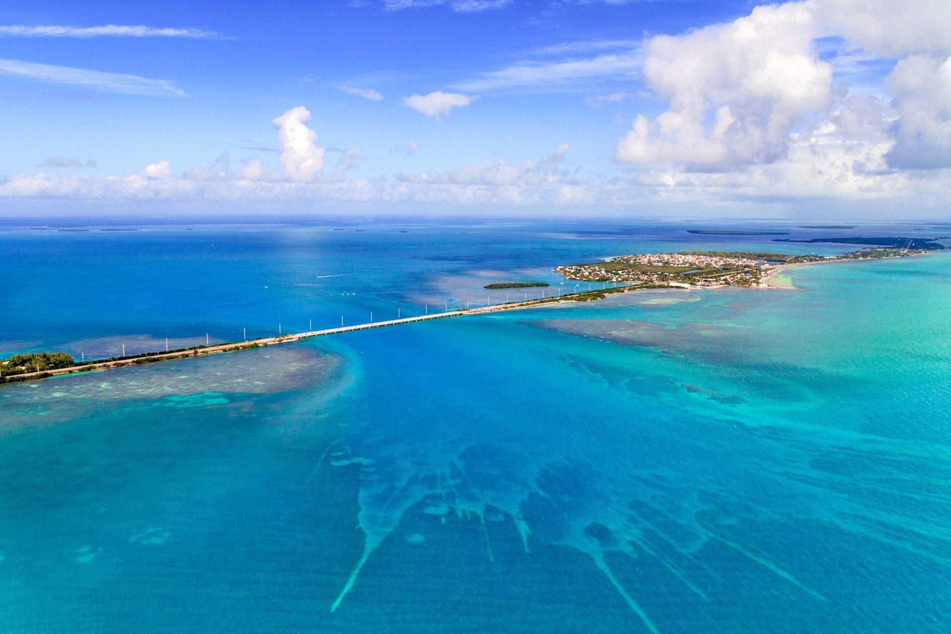 Luchtfoto van een lange brug over turquoise water, die leidt naar een klein eiland onder een blauwe hemel met wolken.