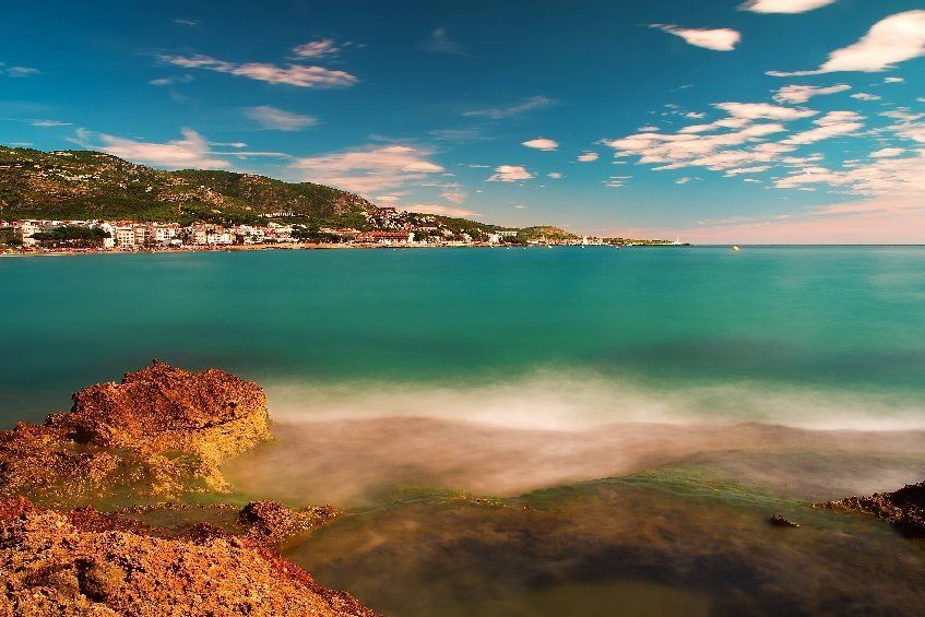 Uitzicht op de kust: turquoise water, wazige golven, rotsachtige voorgrond, dorp op een heuvel onder een blauwe hemel.
